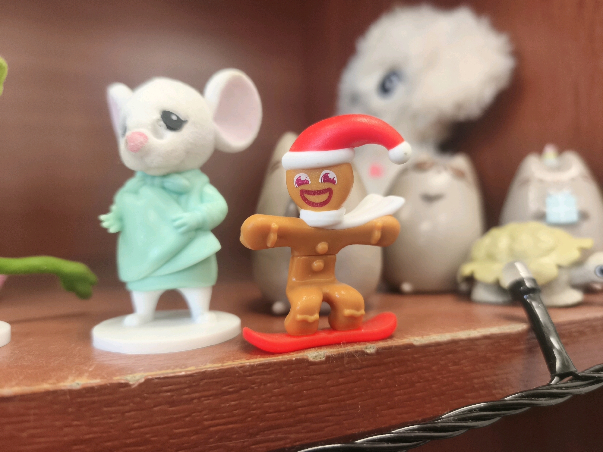 A still life image captures a collection of small figurines arranged on a wooden shelf. The focal point is a gingerbread man, wearing a red Santa hat and a white scarf, positioned on a red snowboard. To the left of the gingerbread man is a miniature white mouse figurine dressed in a teal outfit. In the background, a larger, fluffy white creature with blue eyes is visible, along with several other indistinct figures, including what appears to be a light yellow turtle figurine. The shelf itself has a rough, textured wooden surface with some minor imperfections. The lighting suggests an indoor setting, possibly a display or collection. The image does not contain any elements that would indicate a specific location in the city of Otradnoye, Russia, or any actions or interactions between people.