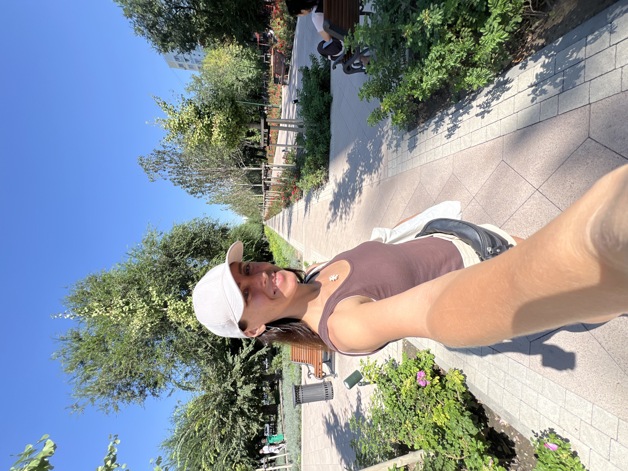A woman wearing a white baseball cap and a brown tank top is taking a selfie in a park. She is smiling and looking at the camera. The park features a paved walkway, benches, trees, and flowerbeds. In the background, there are other people walking and sitting on benches. The sky is clear and blue. The location is identified as Almaty, Kazakhstan.