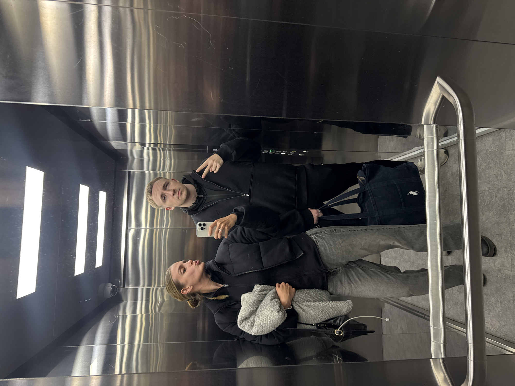 A photograph shows two people taking a selfie in what appears to be an elevator. The setting is a modern elevator with stainless steel walls and bright, linear lighting fixtures. A security camera is visible. The female is wearing a black puffer jacket and light-colored jeans and holding a grey blanket. The male is wearing a black hoodie and pants, and he is making a peace sign. He is also holding a blue bag. The elevator has a metallic handrail on the right side. The floor is gray.