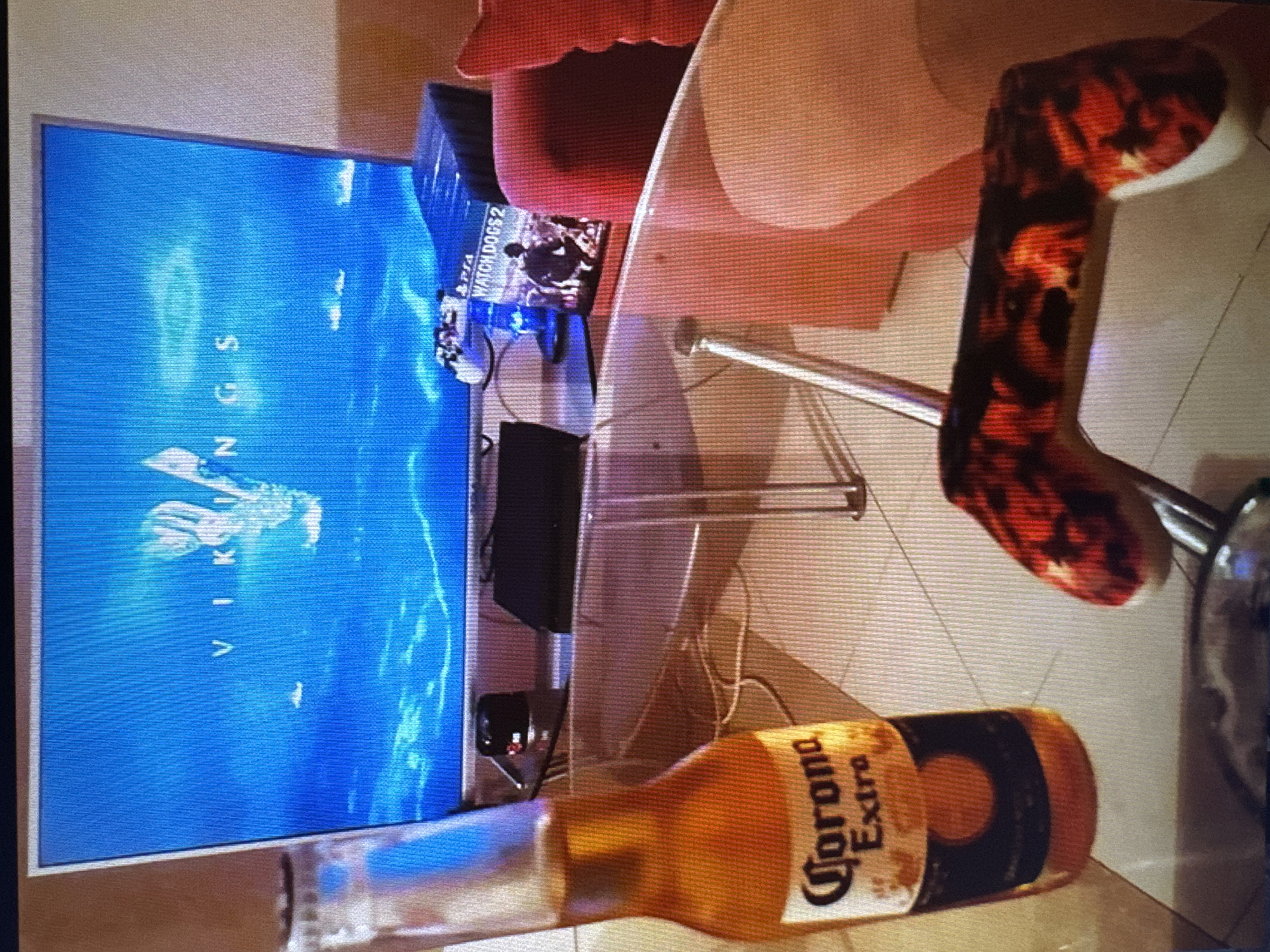 The image shows a glass-topped coffee table in a domestic setting, possibly in the city of Varna, Bulgaria. On the table, there is a bottle of Corona Extra beer, a video game controller with a distinctive red and black pattern, and a PS4 game case for "Watch Dogs 2". Behind the table, a television screen displays the title "VIKINGS" in white lettering against a blue background with an image of a ship. The background shows a couch, suggesting a living room environment. There are no people visible in the image.
