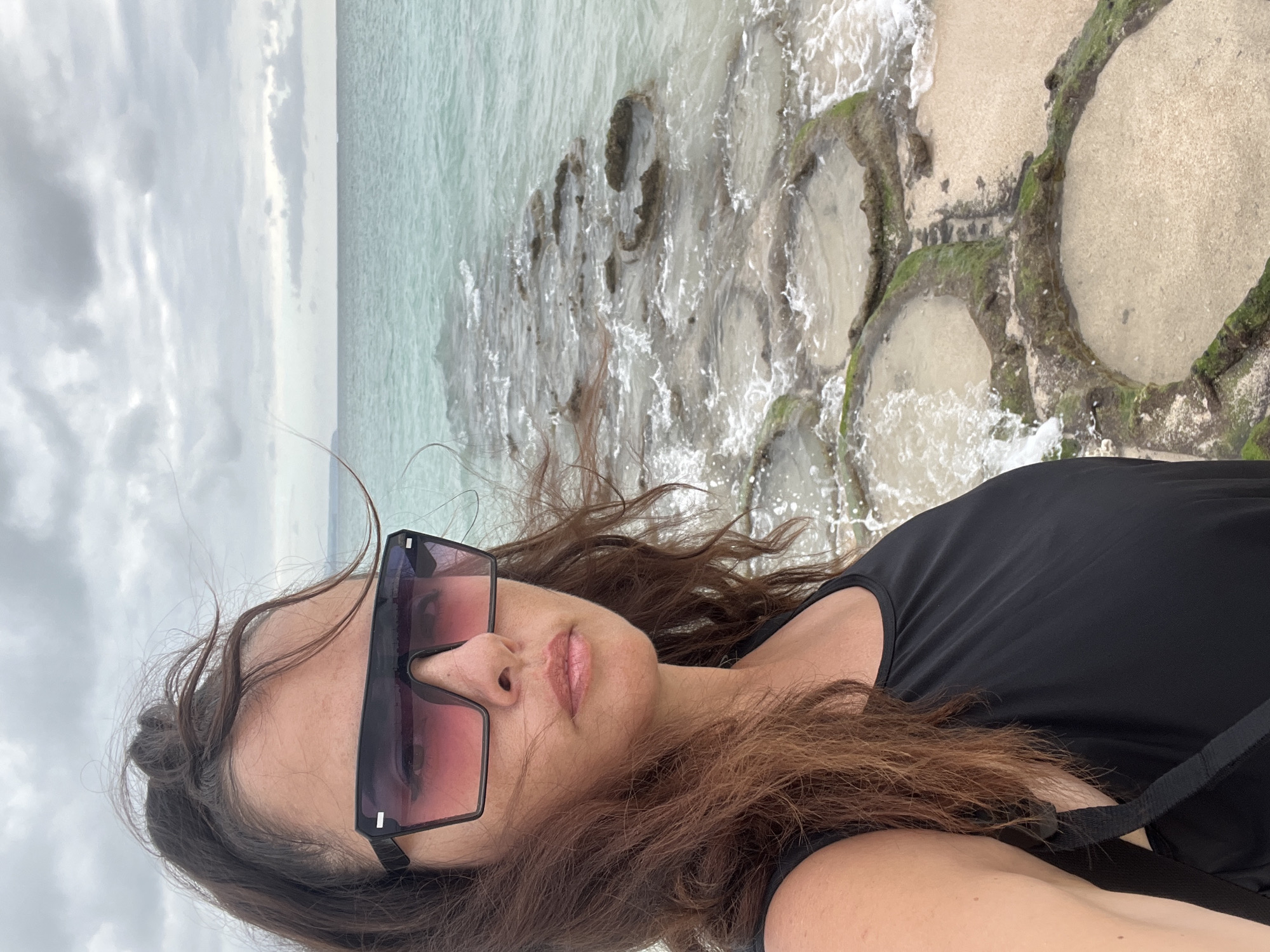 A woman with dark brown hair pulled back in a ponytail and wearing large, dark-rimmed sunglasses with pink gradient lenses takes a selfie on a beach. She is wearing a black tank top. Her head and upper body are in the foreground, with the ocean and a rocky shoreline in the background. The water is a bright turquoise color, and the sky is overcast. The shoreline is made up of natural rock formations that create tidal pools. The scene appears to be located in the city of Kuta, Indonesia, given the distinctive geological formations and the general coastal environment.