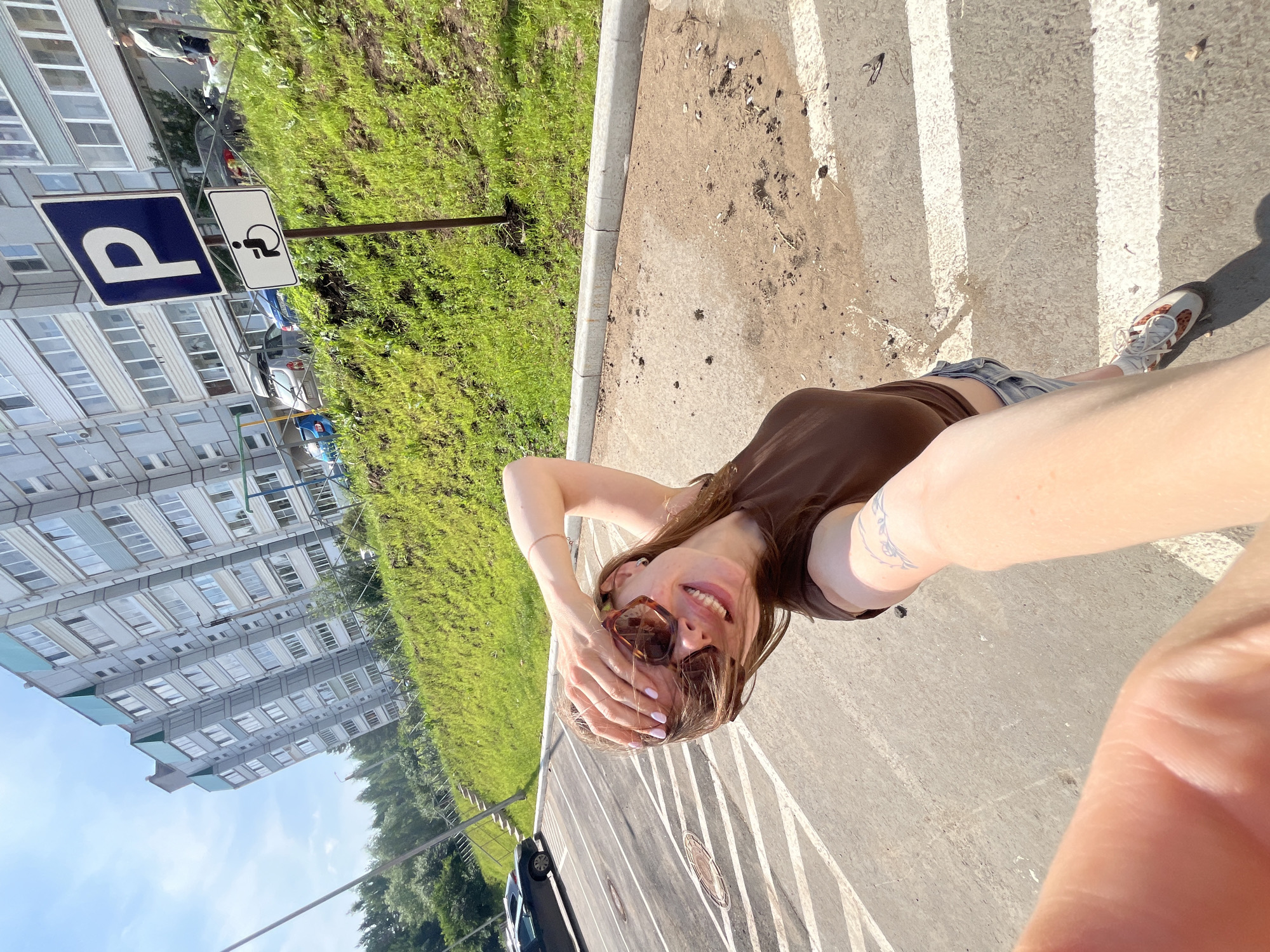 In Chaikovsky, Russia, on a bright, sunny day, a young woman is taking a selfie in a parking lot. She is wearing sunglasses, a brown top, and denim shorts. Her left hand is covering her eyes, and her right arm is extended, holding the camera. A tattoo is visible on her right arm. The parking lot has white lines marking the spaces. Behind her, a grassy embankment slopes upwards, with a signpost featuring a blue "P" for parking and a symbol for disabled parking. Beyond the embankment, a multi-story residential building stands under a clear blue sky. Several cars are parked on the street or in the parking area. Trees line the top of the embankment. The overall scene depicts a typical urban environment in Chaikovsky during fair weather.