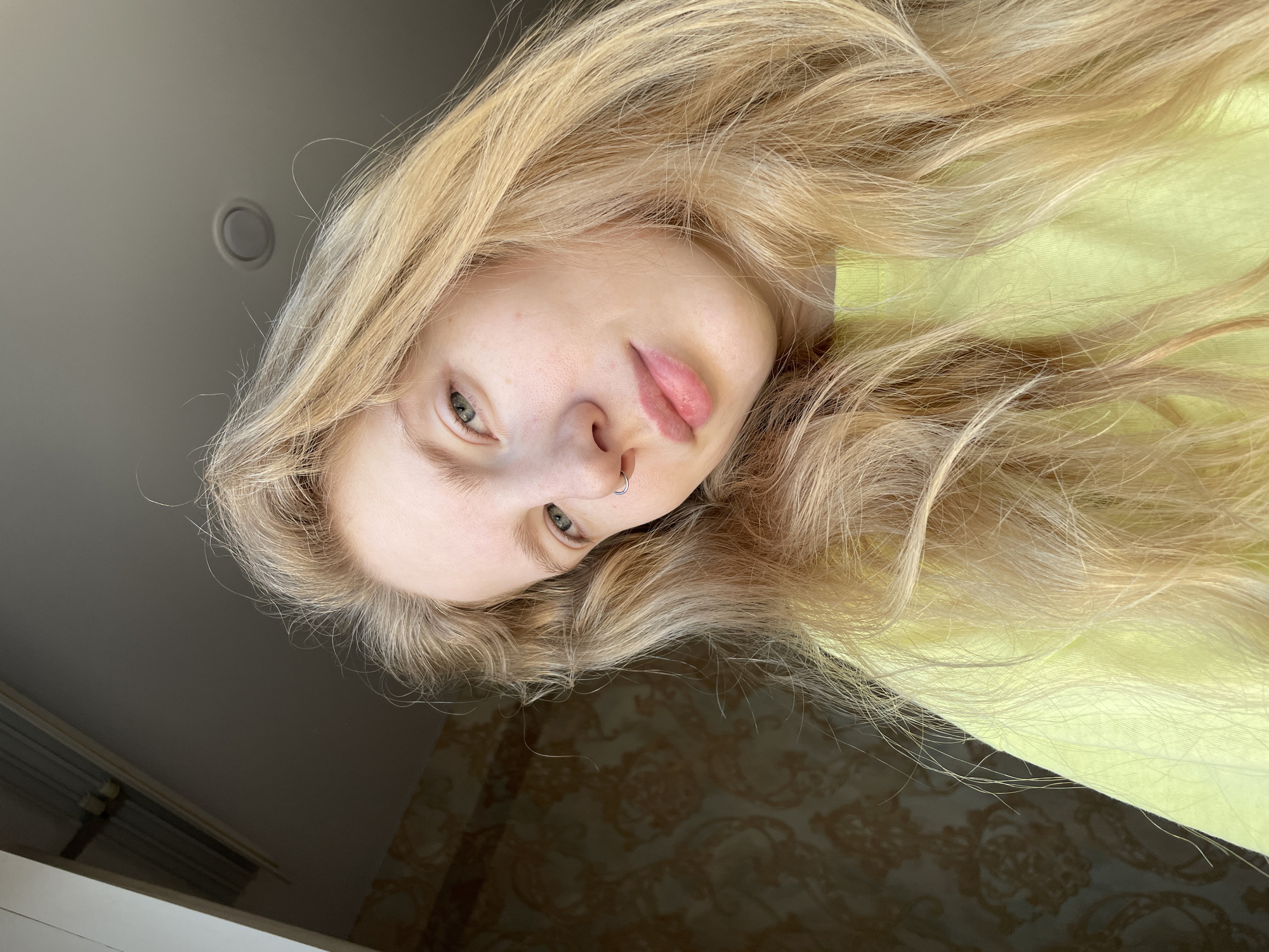 A woman with long, light blonde hair is captured from a low angle. She is fair-skinned and has a nose ring. She is wearing a yellow shirt. The background is a combination of a light gray ceiling with a circular light fixture and a wall with dark brown patterned wallpaper. The location, Izhevsk, Russia, is inferred.
