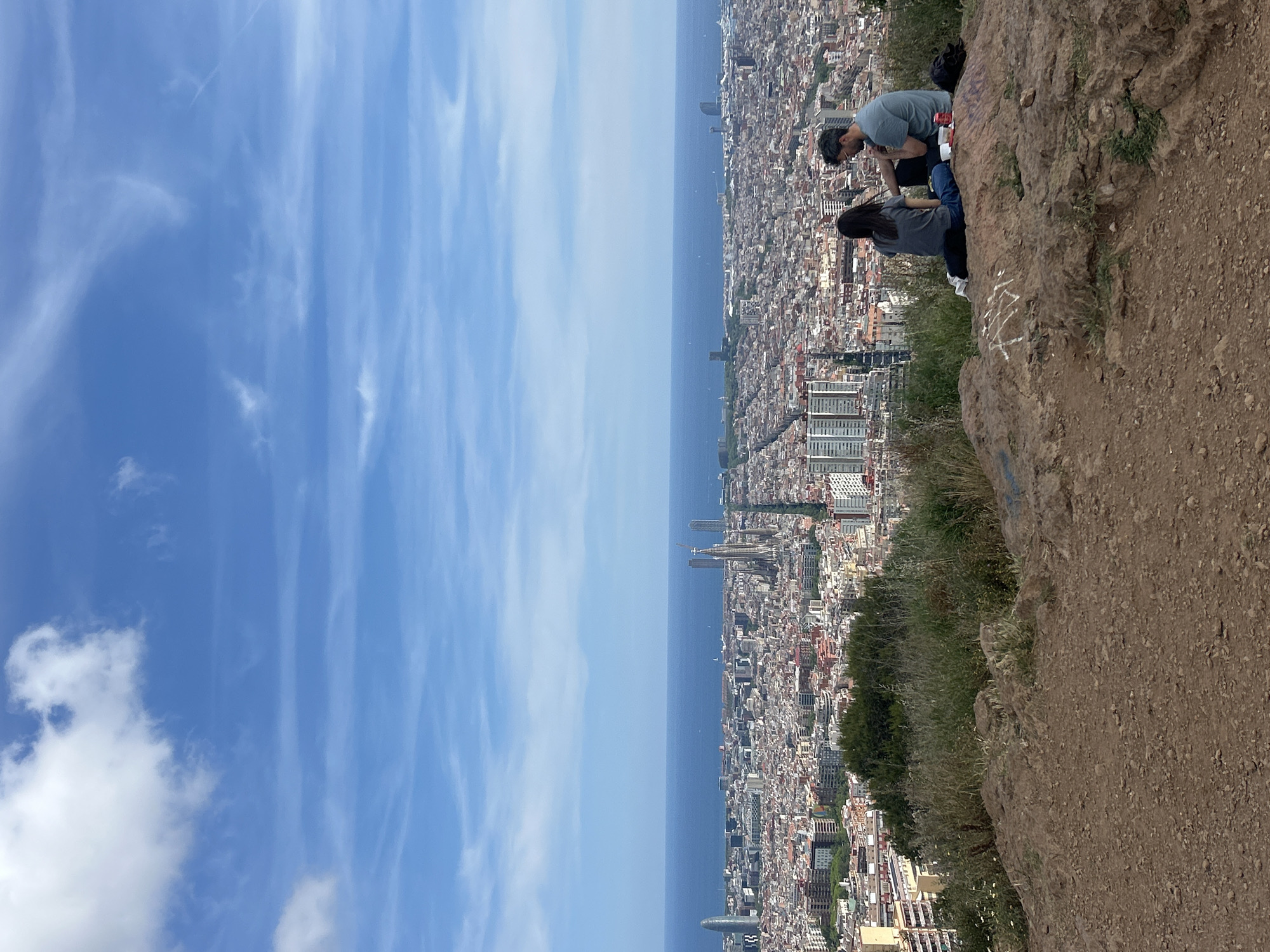Here's a detailed description of the image:
**General Scene:**
The image presents a panoramic view of Barcelona, Spain, observed from a high vantage point, possibly from a hill or mountain. The scene is divided into three main sections: the sky, the cityscape, and the foreground featuring a natural terrain.
**Sky:**
The sky occupies the upper left portion of the image. It is predominantly blue with wispy, elongated clouds scattered throughout.
**Cityscape:**
The majority of the image shows a densely packed urban environment. The buildings are diverse in height and design. Major landmarks can be identified. A body of water (likely the Mediterranean Sea) is visible behind the city.
**Foreground:**
The lower right area is occupied by the edge of the hill or mountain. It features a rocky, dirt-covered ground with some vegetation.
**People:**
Two individuals are positioned near the edge of the vantage point. One appears to be male, the other female. Both are crouched or sitting and observing something below them. Their specific actions or interactions are not entirely clear from the image, but they seem to be looking at the city view.
**Location:**
Given the presence of the Mediterranean Sea and architectural landmarks visible in the cityscape, the location is likely in Barcelona, Spain, from a vantage point on a hill or mountain overlooking the city, possibly Montjuïc.