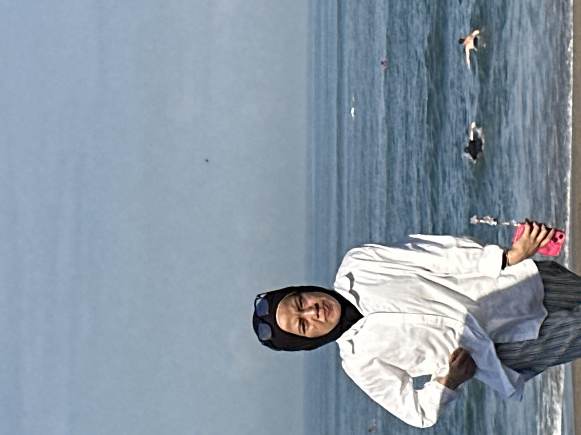 In North Kuta, Indonesia, a woman wearing a black hijab and sunglasses is standing on a beach, facing the camera. She is wearing a white button-down shirt over striped pants and holding a pink smartphone in her right hand. The ocean is visible behind her, with several surfers in the water. The sky is clear and blue.