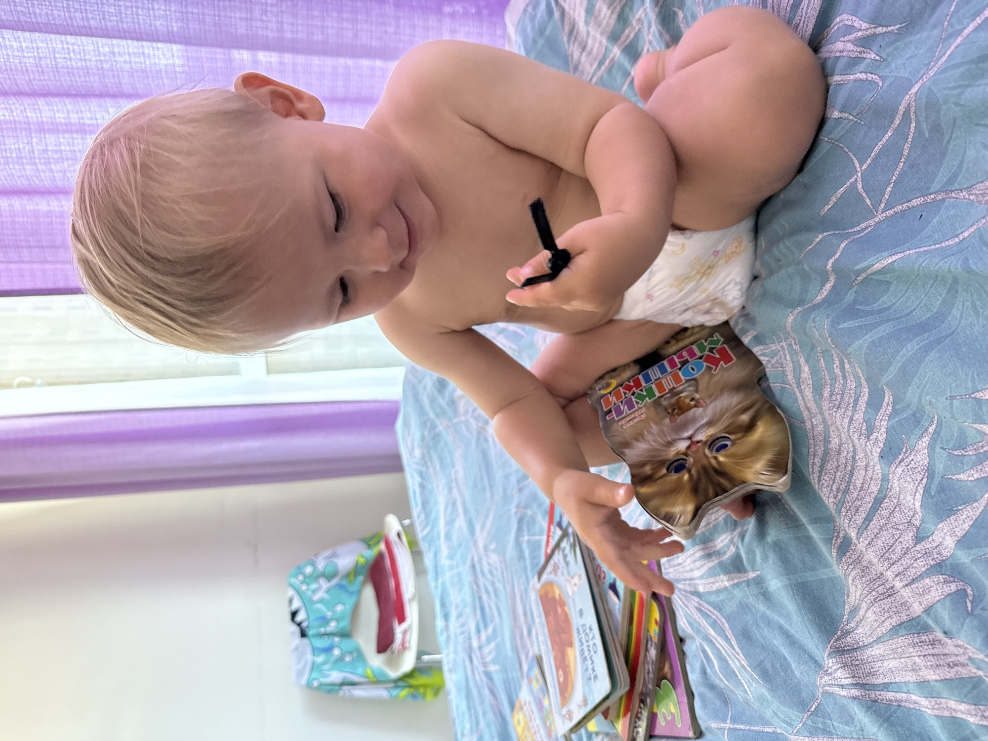 A blond-haired baby is lying on a bed with blue floral sheets, wearing a diaper. The baby is holding a small, shaped book with a picture of a cat on the cover. The baby is looking down at the book and smiling, appearing engaged and happy. In the background, there are purple blinds. To the left of the baby, a colorful cloth organizer with toys is visible. The scene suggests a casual, indoor setting, likely in a child's room or bedroom. The time of day is not explicitly indicated, but the lighting suggests it could be daytime. The location is noted as Sochi, Russia. The book has text on its cover, which appears to be in Russian, with the title partially visible.