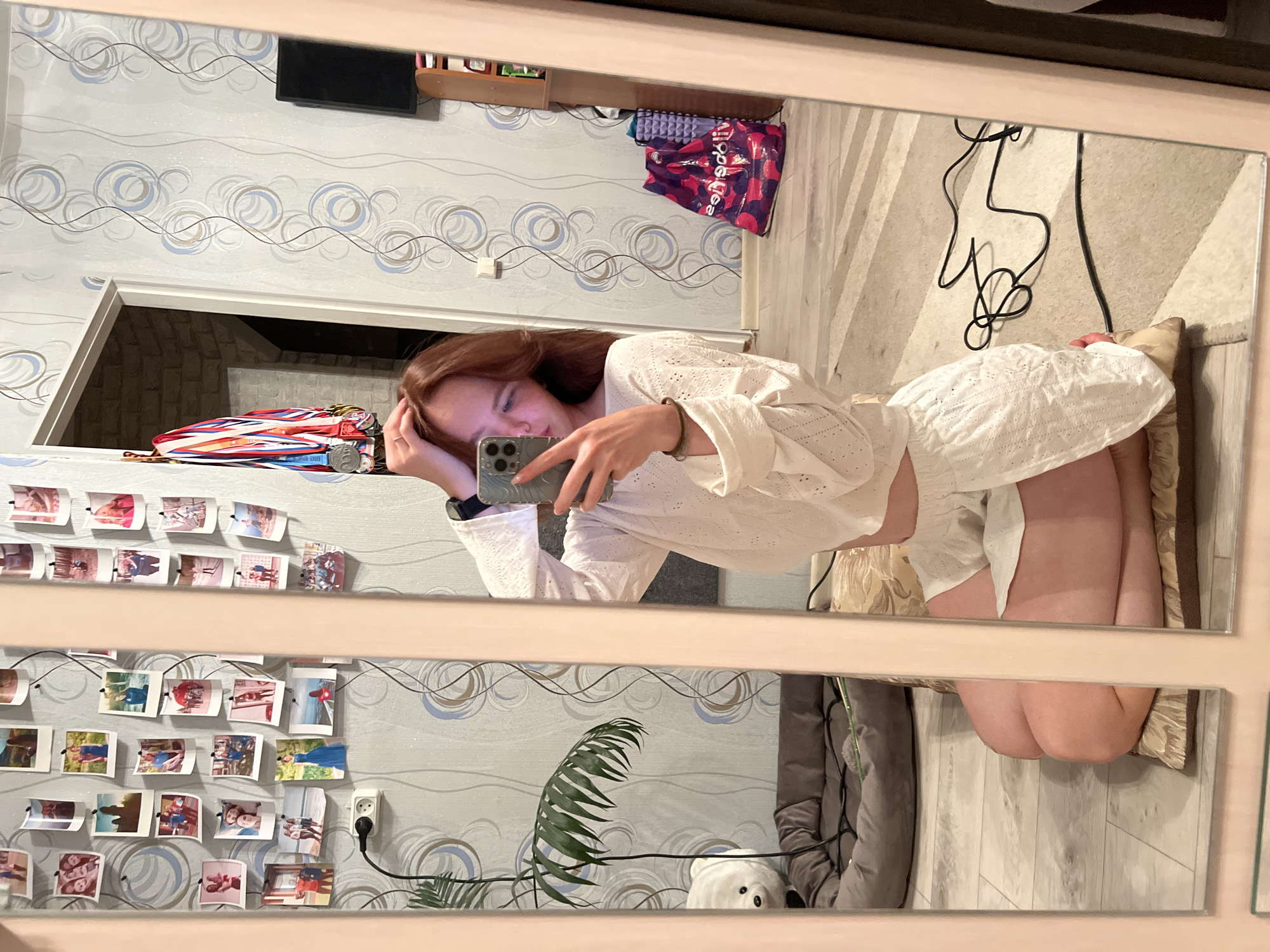 A young woman is taking a selfie in a mirror. She is wearing a white, eyelet top and matching shorts. She is kneeling on a patterned cushion or rug. The background includes a wall with decorative wallpaper featuring concentric circles, a string of photos clipped to the wall, a doorway leading to a darker space, and various items on a shelf above the doorway. A bag with lettering in a Cyrillic script is partially visible. The reflection shows a plant, a stuffed animal, and what appears to be a coat hanging on a hook. Based on the style of the room and the Cyrillic writing, the location is likely Chaikovsky, Russia.