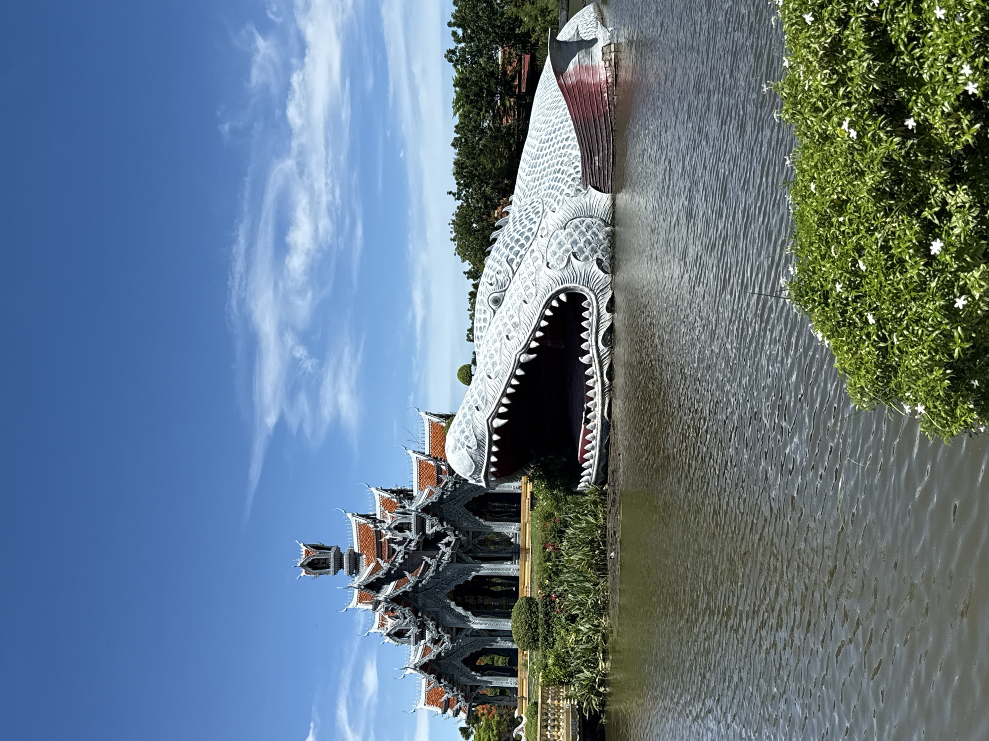 The image captures a distinctive architectural scene in Bang Pu Subdistrict Municipality, Thailand. A predominantly gray and white traditional Thai building with ornate roof structures is visible on the left. The building's intricate detailing is partially obscured by greenery. The building is either next to or in a body of water. Adjacent to the water stands a large sculpture of a dragon's head with an open mouth. The sky is a clear blue with scattered white clouds.