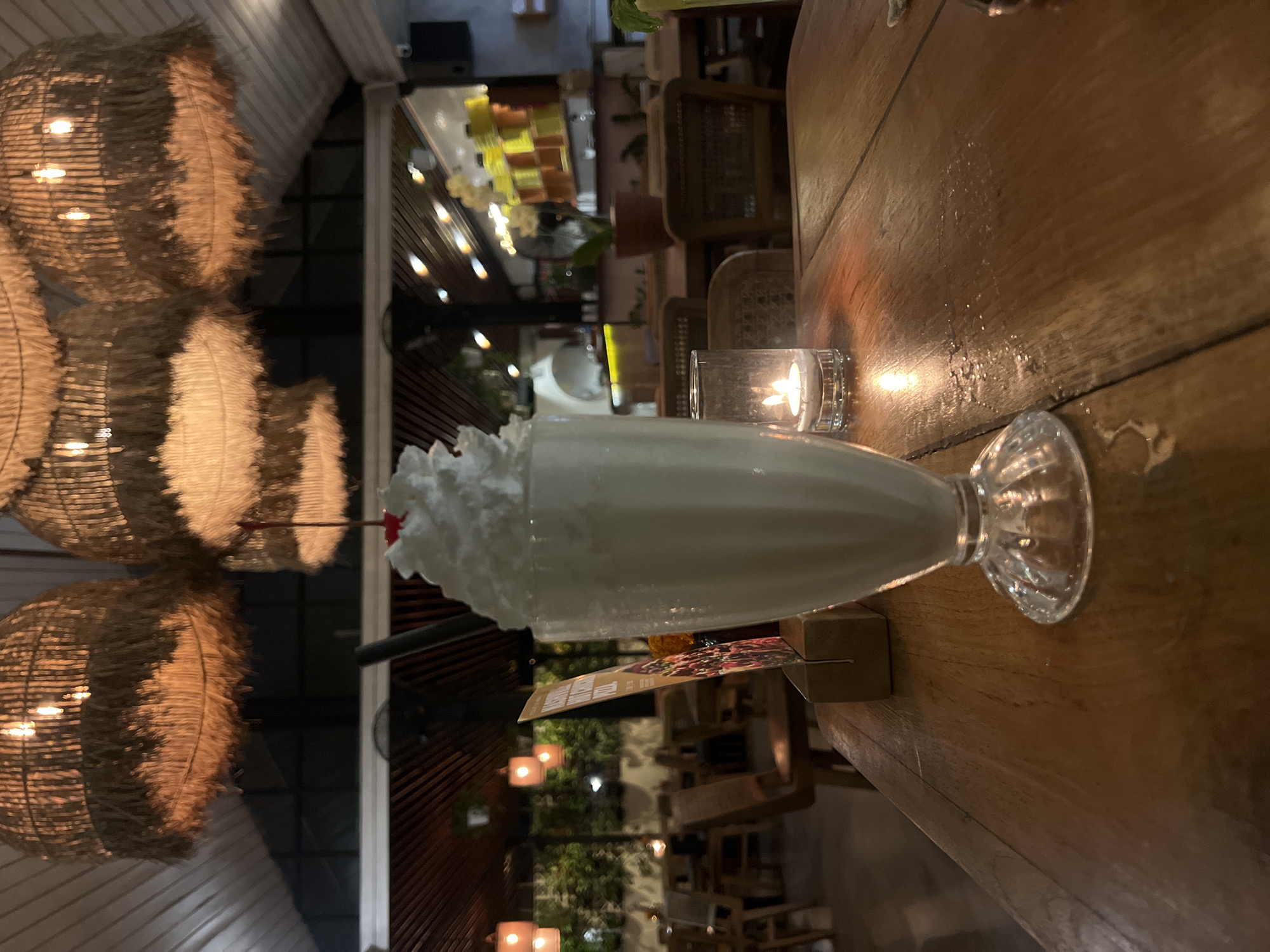 The image shows a vanilla milkshake with whipped cream and a cherry in a clear glass on a wooden table in what appears to be a restaurant or cafe. A small candle in a glass holder is nearby, casting a warm light on the table. The background reveals a blurred outdoor dining area with decorative lighting and possible foliage, suggesting an open-air or semi-outdoor setting. The general ambiance, including the lighting and decor, indicates a casual or upscale casual dining experience. The location in the city of North Kuta, Indonesia is not confirmed by the image alone.