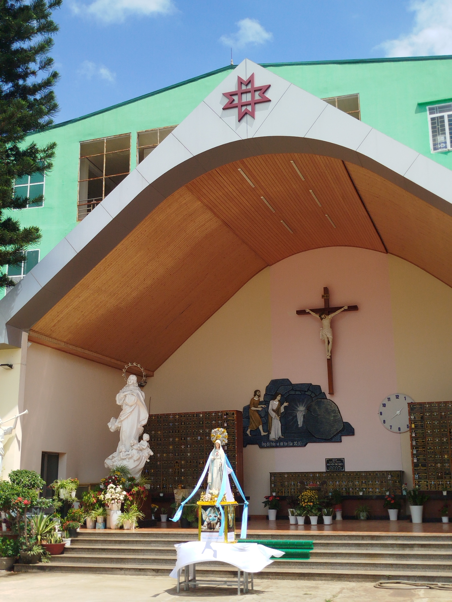 The image shows the facade of a church in Pleiku Ward, Vietnam. The building is painted a light green, with a gabled portico featuring a red star-like design at the peak. The underside of the portico is finished with wooden slats.
Within the portico, several religious statues are visible, including a statue of Mary draped with a blue sash and a statue of Jesus on a cross. Other religious artworks and a large clock are mounted on the wall.
In front of the steps leading to the portico, a table covered with a white cloth is set up, likely for a religious ceremony or event. Various potted plants and flowers are arranged on the steps and around the statues, adding color to the scene. A tree is visible to the side.