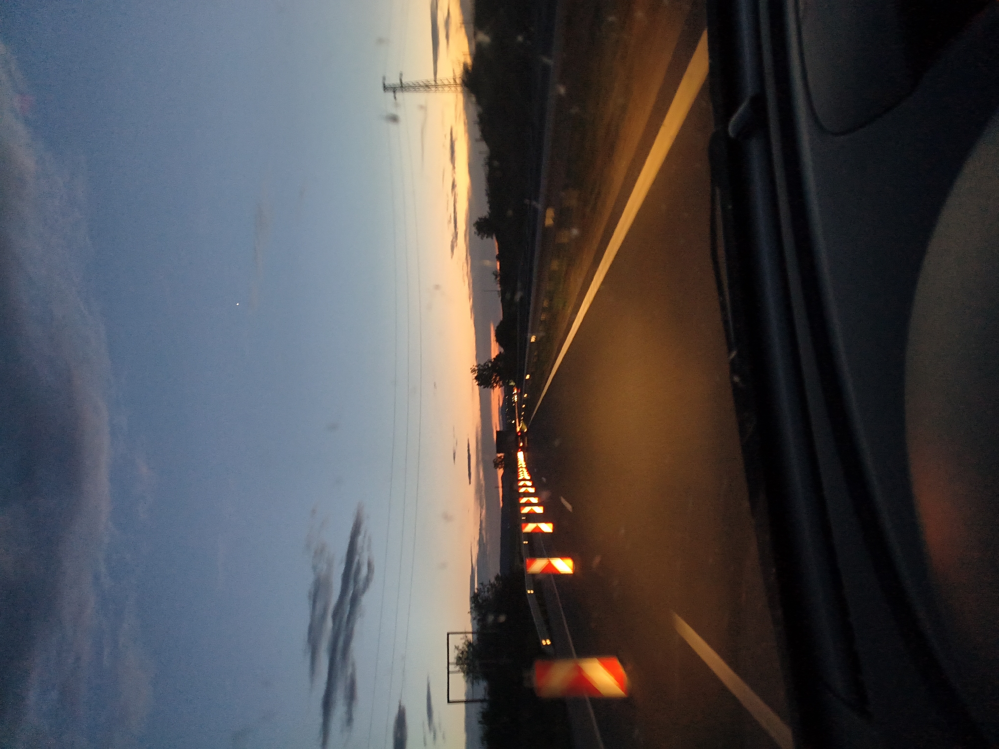 The image, captured from inside a vehicle, depicts a road at dusk, with the sky transitioning from blue to orange and pink near the horizon. A line of hazard markers, in red and white, stretches along the road indicating a construction or hazard zone. Beyond the hazard markers, the road extends into the distance, where there are lights of other vehicles. Power lines are visible against the sky. The interior of the vehicle is visible on the right side of the frame. The location is assumed to be in or near the city of Shumen, Bulgaria.