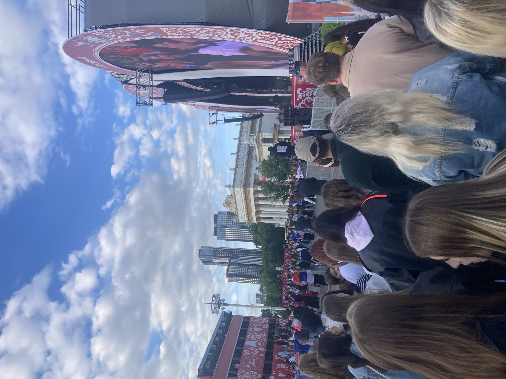 The image depicts a large outdoor public event, likely a concert or festival, taking place in Moscow, Russia, during daytime. The view is from behind a large crowd of people, mostly seen from the back, all facing a prominent circular stage.
The stage features a large LED screen displaying moving images, and its outer edge is adorned with a decorative red border incorporating white patterns. Part of a Russian word, "БЕДА", is visible on this border, strongly suggesting the word "ПОБЕДА" (Pobeda, meaning Victory). Sound equipment and scaffolding are visible around the stage structure.
In the mid-ground, behind the stage, a grand, classical-style building with ornate columns and sculptures stands out, characteristic of structures found at VDNKh (Exhibition of Achievements of National Economy) in Moscow. Further in the background, modern skyscrapers rise against the horizon. To the left of the frame, a red building or structure with white decorative patterns, similar to those on the stage, is partially visible.
The weather is clear with a bright blue sky and scattered white clouds. The natural lighting indicates it is either late morning or afternoon. The overall scene conveys a sense of a large, lively public gathering.
