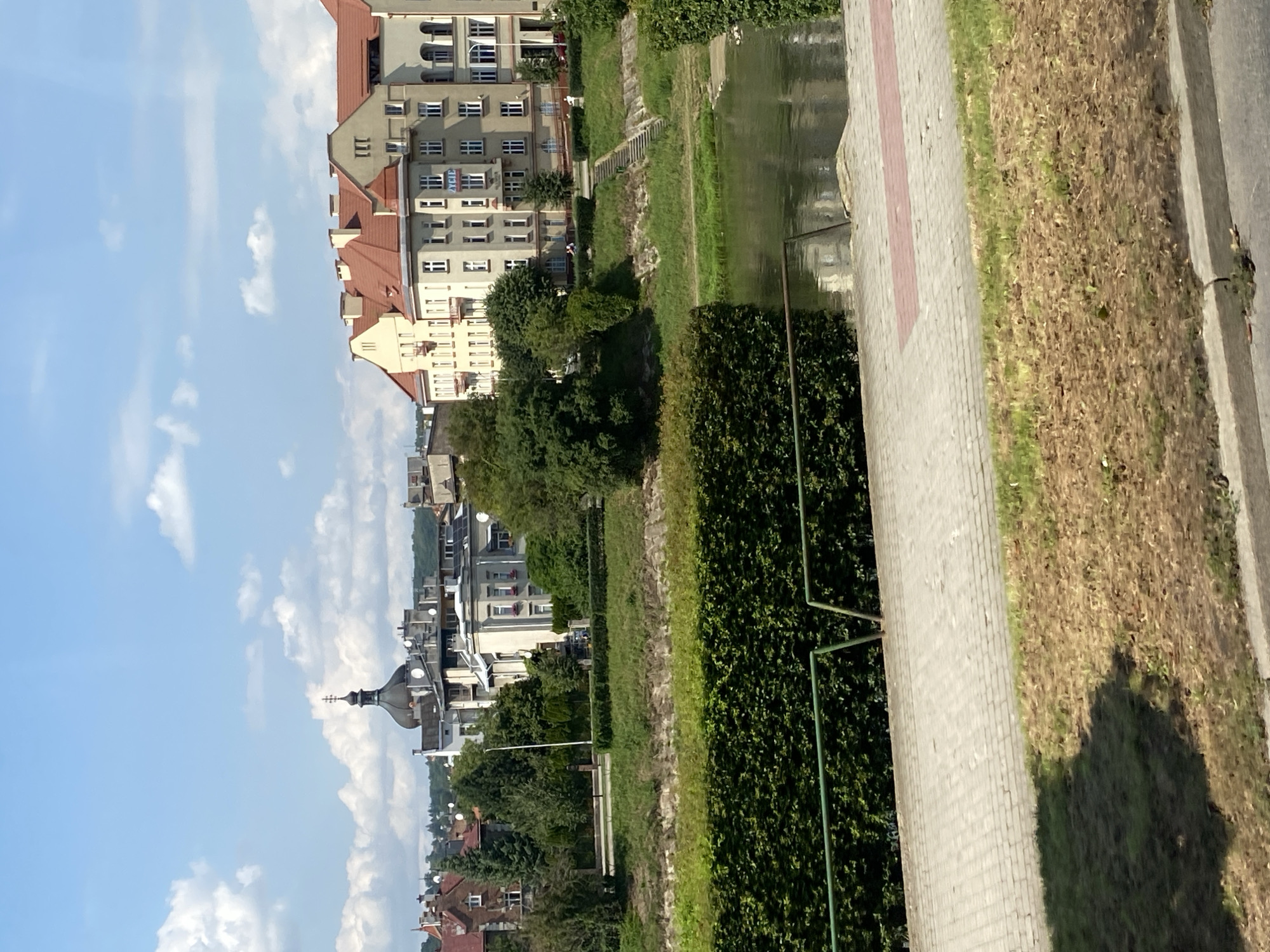 The image depicts a riverside scene in the city of Przemyśl, Poland, on a clear, sunny day with scattered clouds. In the foreground, a paved walkway runs parallel to a lush green hedge that separates it from a body of water, likely the San River. Beyond the hedge and water, a grassy embankment rises, leading to a row of buildings with red-tiled roofs. The buildings are varied in style and architecture, with some featuring classical elements and others more modern designs. One building displays a sign with Polish text, reading "ŻYCIE". Further in the background, a distinctive church dome with a cross is visible, as are other structures and trees, suggesting an urban setting. The overall impression is one of a tranquil city view, with nature and architecture coexisting along the river.