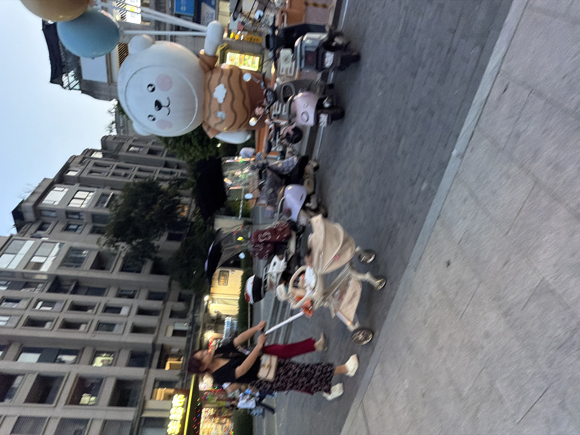 The image depicts a street scene in Jiangjin, China, with a focus on a woman pushing a baby stroller. She is wearing a black top, a patterned skirt, and light-colored shoes. To her right, a person in red pants walks alongside. In the background, several parked motorbikes, some with umbrellas attached, are visible, suggesting a transportation hub or a marketplace. A large inflatable cartoon character, resembling a bear, stands prominently in the middle ground, with two balloons above it. The buildings in the background are multi-story residential or commercial structures. The overall lighting suggests it is either dusk or dawn.
