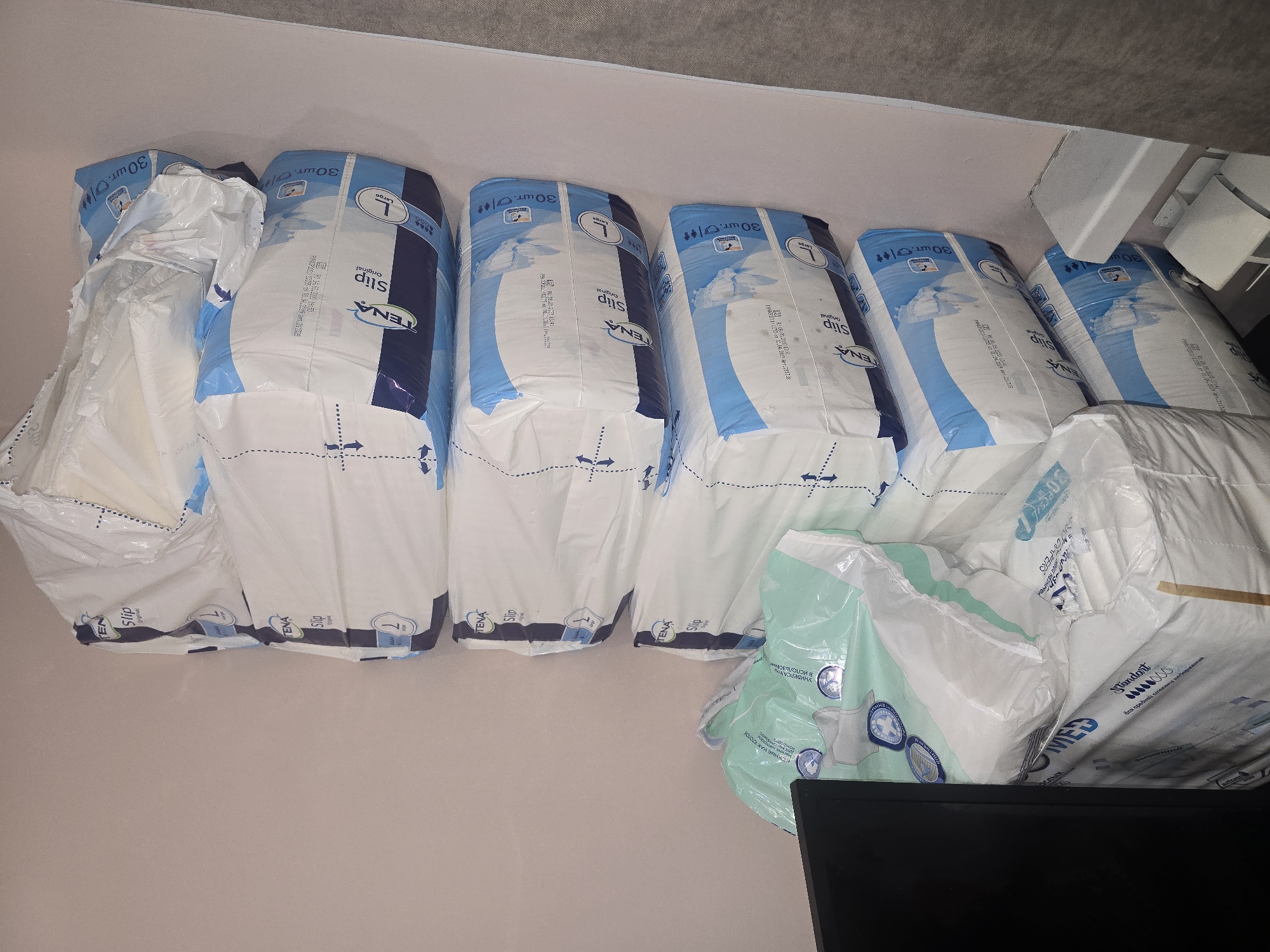 The image depicts a corner of a room with a stack of adult diapers against a wall. The diapers are packaged in plastic wrapping, with several brands visible including "TENA Slip Original" and "MED Standard." The lighting suggests an indoor setting, possibly a home or storage area. No people or specific landmarks of Ulan-Ude, Russia are visible in the image.