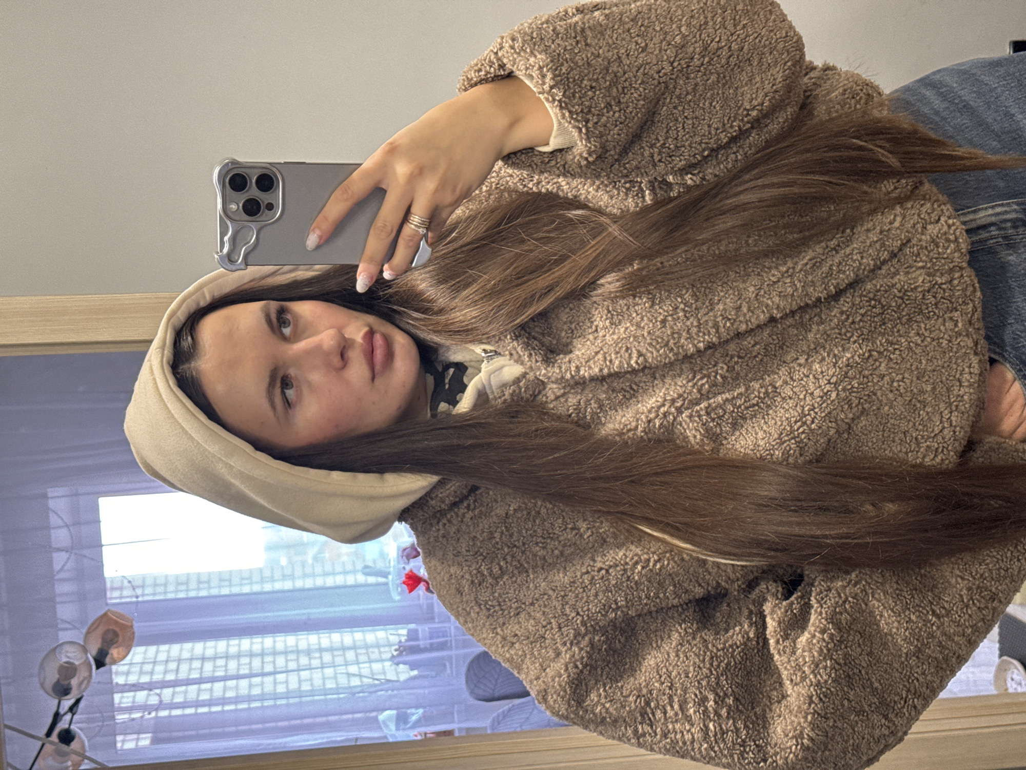 A young woman is captured in a mirror selfie, holding a smartphone. She has long, straight, rich brown hair that cascades over her chest and shoulders. She is wearing a plush, brown, textured hooded jacket, possibly made of sherpa or fleece material, over a light-colored garment. Blue denim jeans are visible on her lower body.
Her face is visible, with a neutral to slightly contemplative expression as she looks into the mirror/phone camera. She has defined eyebrows, natural-looking eyelashes, and full lips. Some minor skin blemishes are visible on her cheeks and forehead.
She holds a grey smartphone with a prominent triple-lens camera module in her right hand. The phone is encased in a distinctively shaped grey protective case with an intricate, somewhat abstract design on the side. Multiple rings adorn the fingers of her right hand, and her nails feature a light, patterned manicure.
The background, reflected in the mirror, reveals an indoor setting. A window with horizontal blinds or slats is visible, allowing natural light into the space, suggesting it is daytime. A decorative lamp with multiple round, possibly amber-toned bulbs is also reflected. The room features a light-colored wall and a wooden frame or ledge.
There is no visible text in the image.