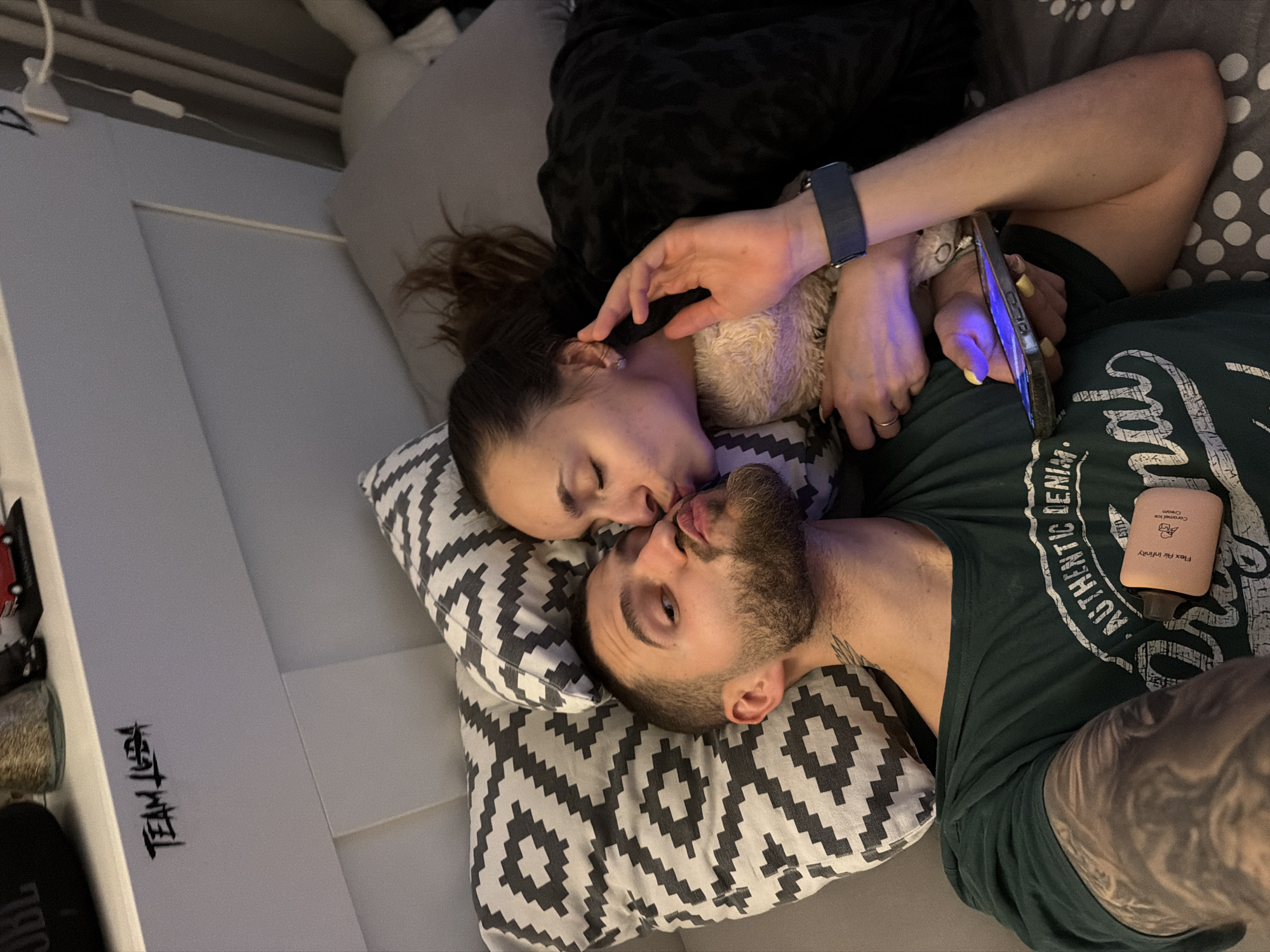 A high-angle shot depicts a couple lying on a bed. The woman, with dark hair pulled back, is on her side facing the man. The man, with a beard, is on his back. They are kissing. The woman has her arm around a stuffed animal. In his hand is a phone with a blue screen. The man wears a dark green t-shirt. Visible next to him is a bottle of "Flex Air Infinity" cream. A pillow with a black and white geometric pattern is under their heads. The location appears to be indoors. It is not possible to verify that the scene is taking place in Sofia, Bulgaria.