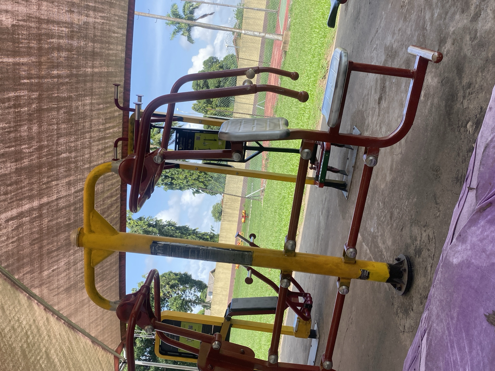 The image shows a piece of outdoor exercise equipment in a public park in Ikorodu, Nigeria. The equipment is primarily yellow and red, with silver seats and hand grips. The park has a grassy field, trees, and a running track. The area is sheltered by a canopy, and there are residential buildings in the background.