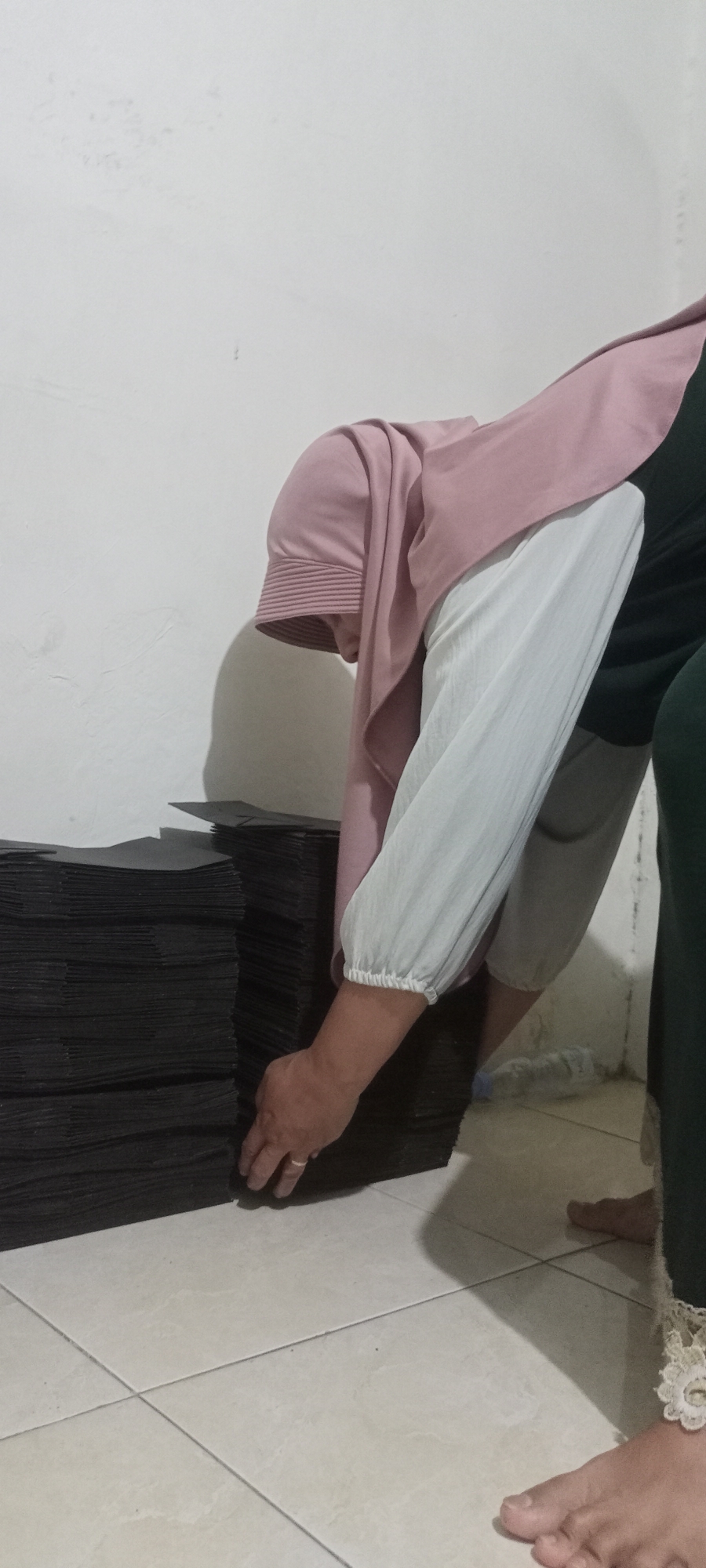 A person wearing a pink hijab and a light-colored long-sleeved shirt is bending over to stack black, flat objects in front of a white, textured wall. The person's right hand is extended towards the stack of black objects, which appear to be some form of paper or cardboard, organized in two large piles. The person is also wearing light-colored trousers and is barefoot. Another person is partially visible on the right side of the frame, also barefoot and wearing dark green trousers with lace trim. The scene appears to be indoors, with a tiled floor and a slight shadow cast on the wall behind the person bending over. The location is identified as Gempolklutuk, Indonesia.