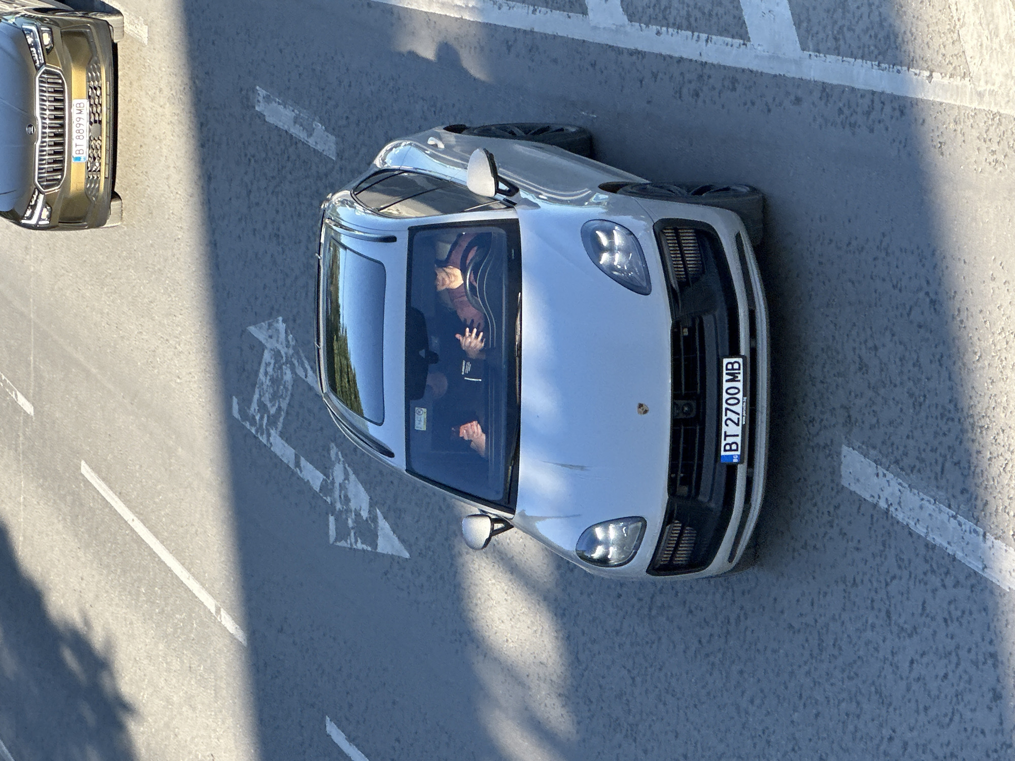 An aerial view depicts two cars parked on a grey asphalt road with white lane markings. One car, located on the left, is a gold-colored Kia, with the license plate “BT 8899 MB”. The other car, a light grey Porsche with license plate “BT 2700 MB”, is positioned closer to the center of the frame. Two individuals are visible inside the Porsche. The location is in Veliko Tarnovo, Bulgaria.