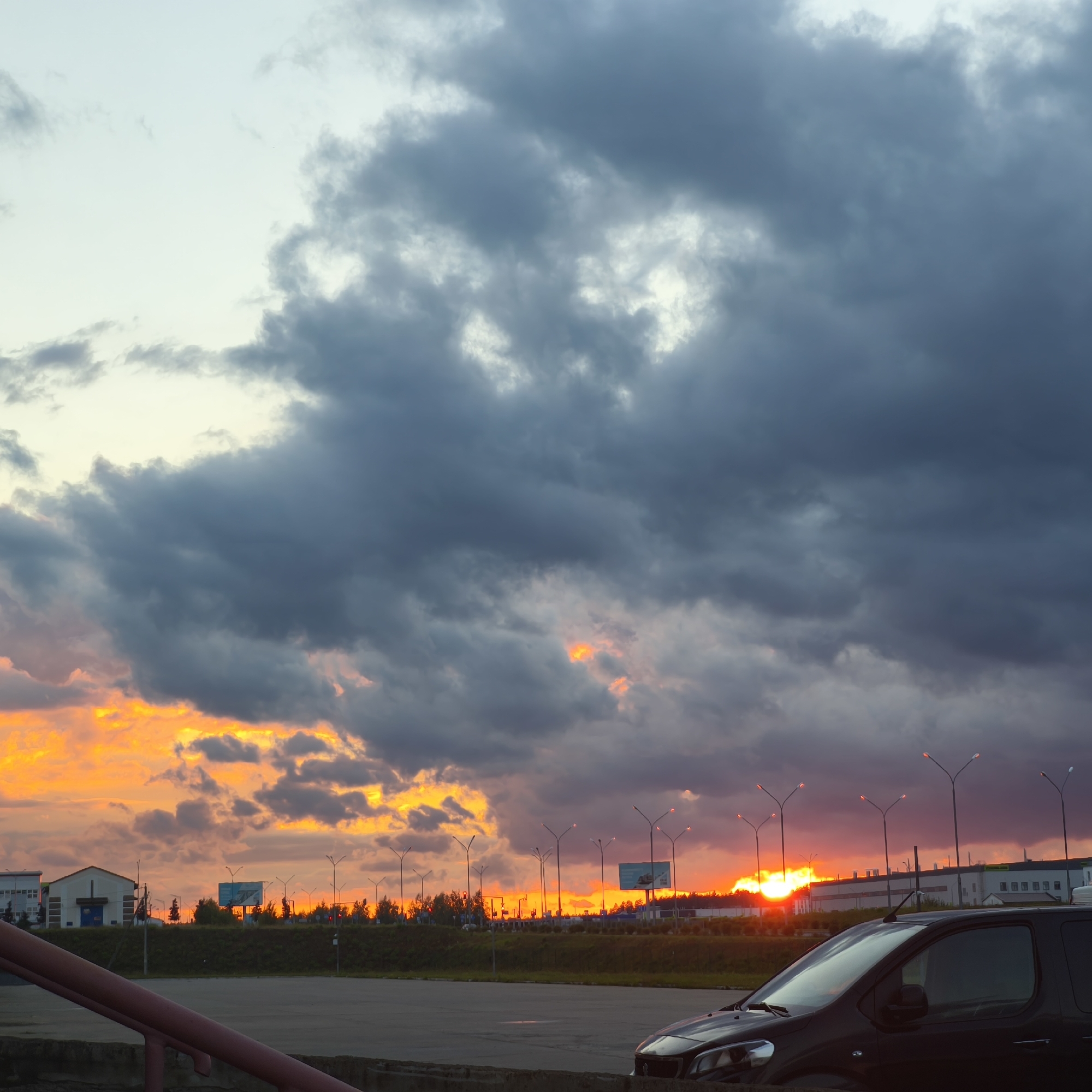 A high-angle shot captures a twilight scene in the Драчкаўскі сельскі Савет, Belarus, featuring a dramatic sky filled with dark, heavy clouds interspersed with vibrant orange and yellow hues, indicating a sunset. The clouds dominate the upper portion of the frame, creating a sense of impending weather. Below the turbulent sky, a low horizon line is visible, populated by silhouetted trees and industrial or commercial buildings. Streetlights with glowing red-orange tops are scattered across the landscape, their silhouettes sharp against the fading light. In the foreground, a dark-colored vehicle, possibly a van or SUV, is partially visible on the right side, facing left. The vehicle is parked in what appears to be a paved area, possibly a parking lot. A red railing or barrier is in the lower left corner, adding a foreground element. The overall atmosphere is one of a dramatic natural display over a developed, yet perhaps sparsely populated, area.