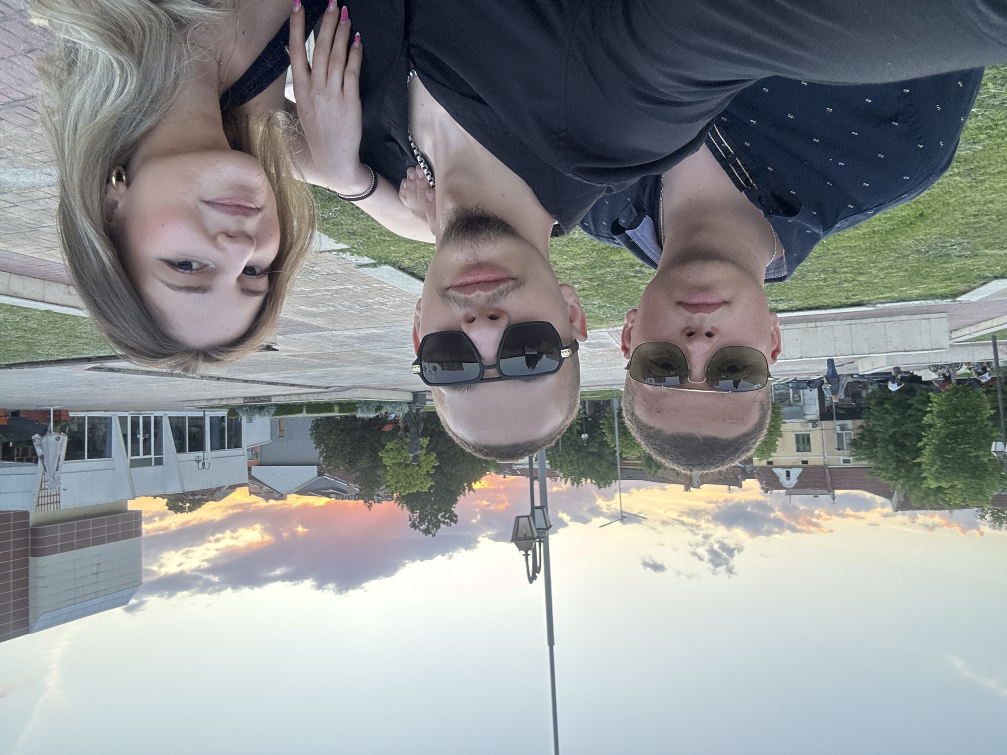Three people are posing for a picture outdoors. A woman with blonde hair is to the left. Two men wearing sunglasses are standing to the right of her. In the background is a building and a sunset sky. This scene is in Pazardzhik, Bulgaria.