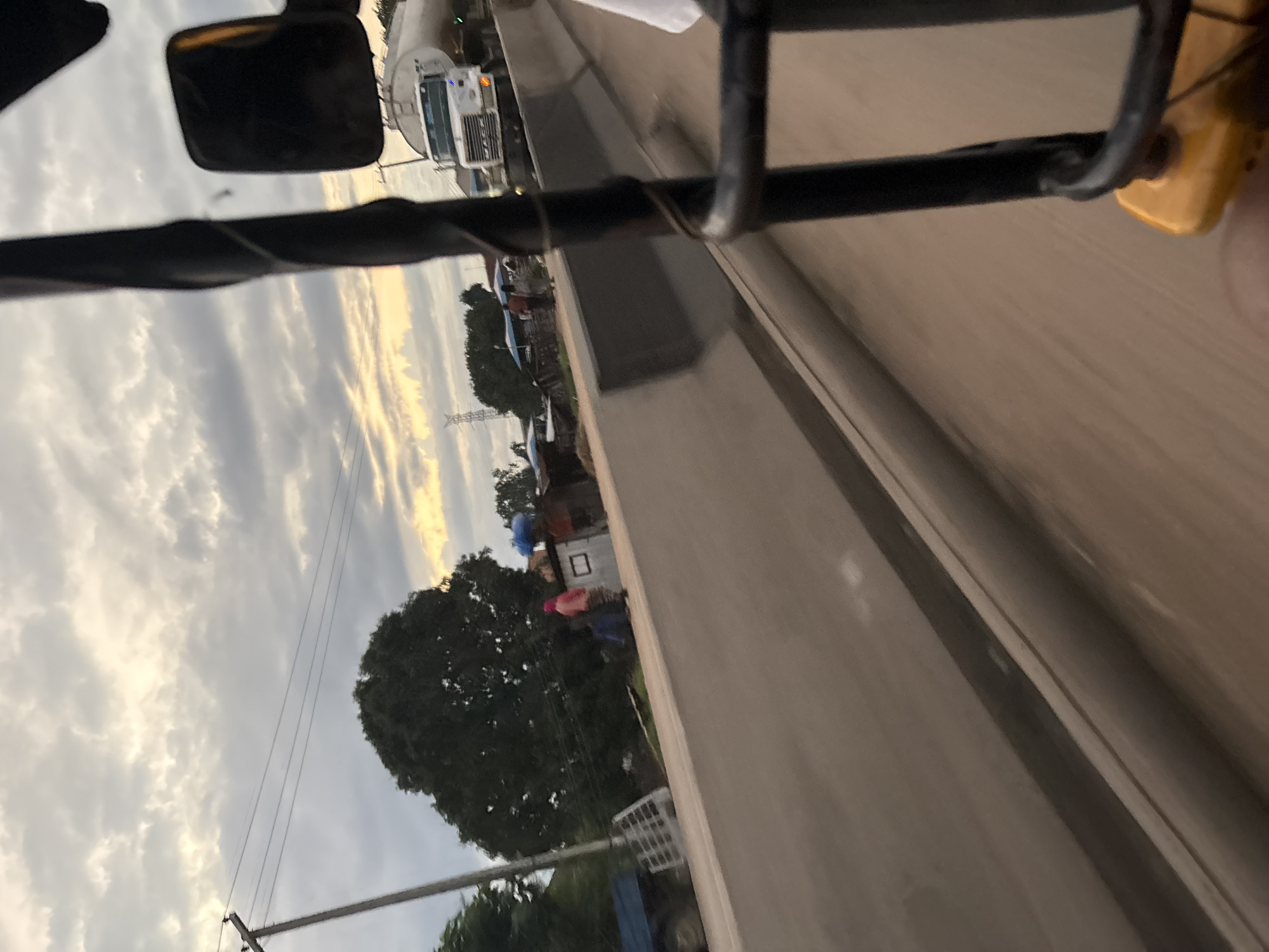 The image depicts a view from a moving vehicle, likely a tricycle or tuk-tuk, in Onitsha, Nigeria. The foreground is dominated by the vehicle's black metal frame and a yellow plastic component. The road surface is visible, appearing to be a light brown, dusty material. A concrete barrier runs along the side of the road. Beyond the barrier, there are some structures, including small buildings and what appears to be a blue truck. A large, dark green tree is situated behind the barrier, with some people dressed in light-colored clothing standing near the structures. In the background, a white tanker truck is visible on a road or highway, and further in the distance, power lines and a large transmission tower can be seen. The sky is overcast with a mix of gray clouds and patches of yellow sunlight, suggesting either dawn or dusk. The overall impression is that of a busy, developing urban or peri-urban environment.