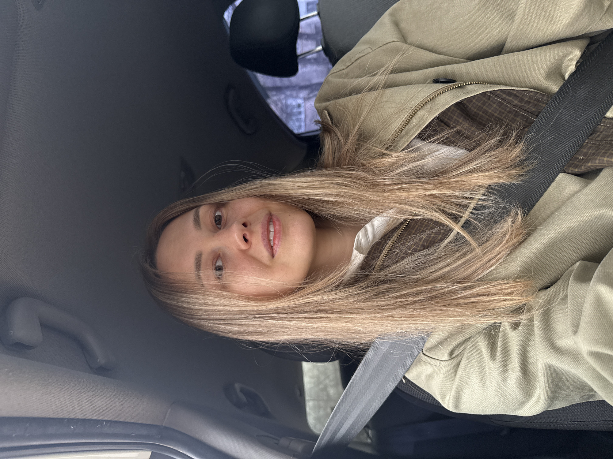 A young woman with light brown hair is wearing a tan jacket and a seatbelt while inside a car. The location is not discernible as Chaikovsky, Russia or any other specific location without additional visual context. The background seen through the car window appears to be an urban environment with trees and buildings. The woman is looking at the camera.