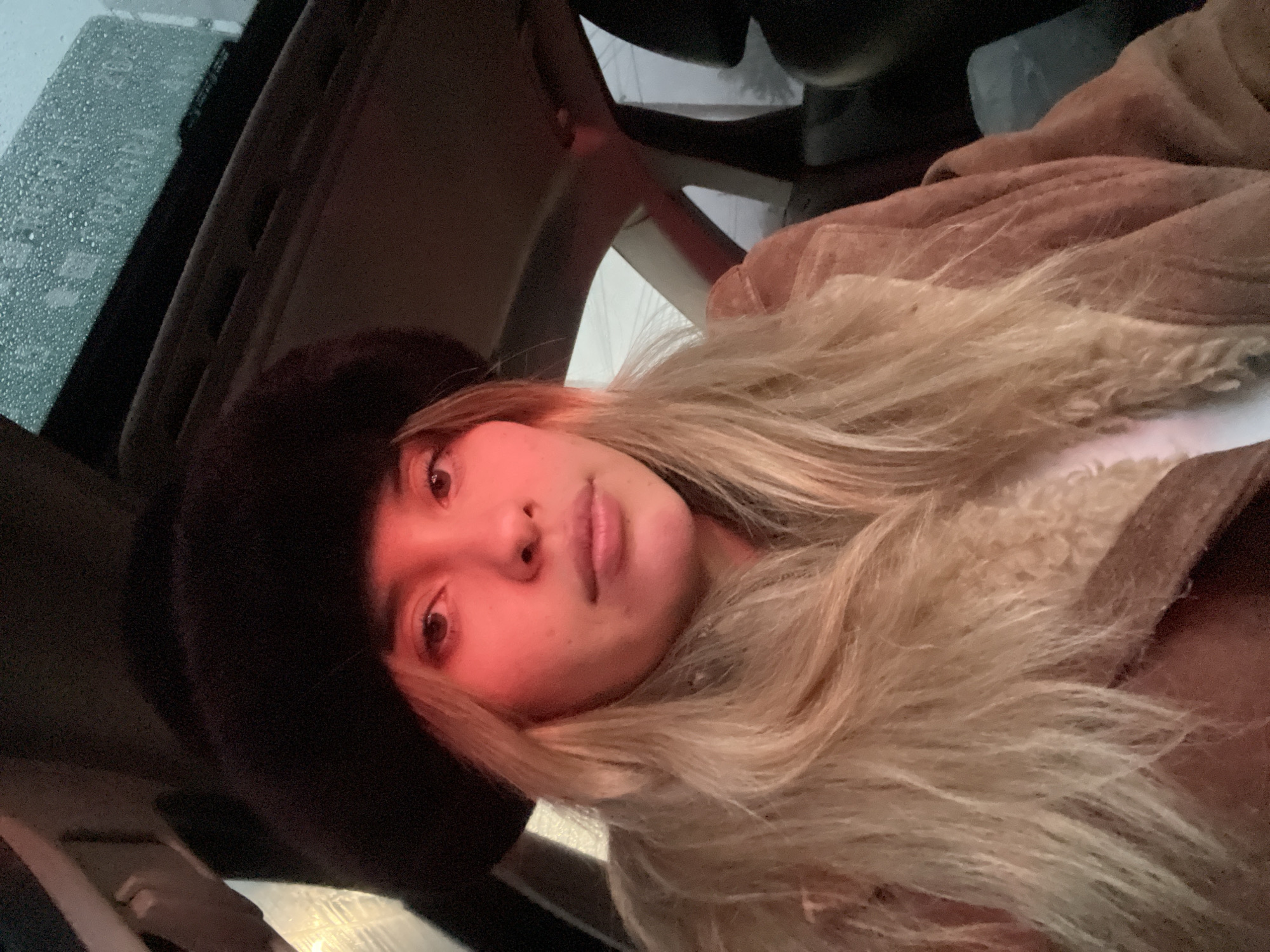 A young woman is positioned inside a car, visible through the sunroof, which shows raindrops. She's wearing a dark furry hat and a light brown, possibly sheepskin, jacket. Her long blonde hair is loosely flowing. The scene is illuminated with a reddish hue. Given the request for a location in San Fernando del Valle de Catamarca, Argentina, without other indicators, I cannot confirm whether this photograph was taken there. I cannot provide a forensic description without additional information. The subject is looking at the camera.
