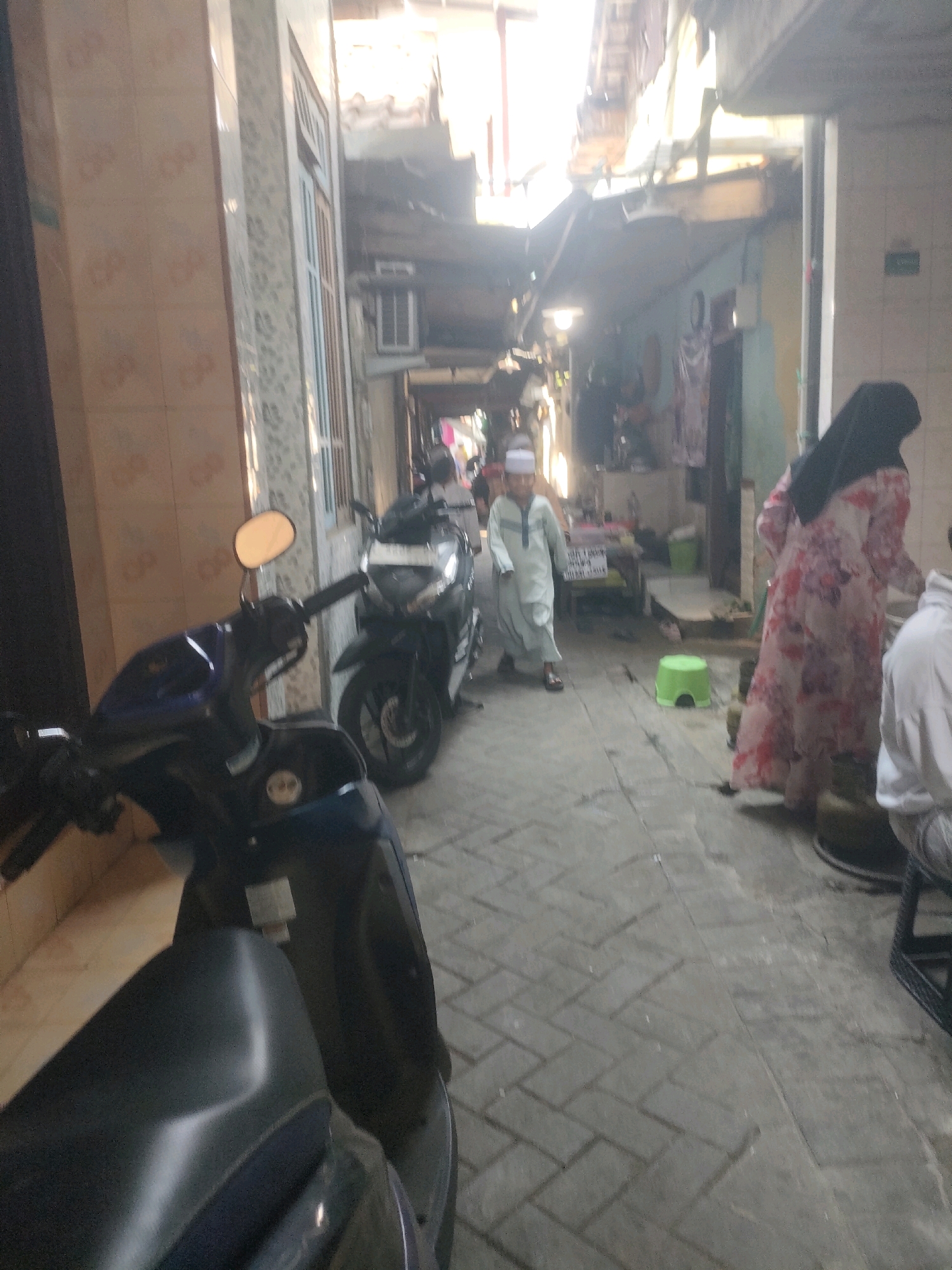 This image depicts a narrow alleyway in Surabaya, Indonesia, likely a residential or commercial area. Two scooters are parked in the foreground on the left side of the frame. A young boy wearing a white cap and a light blue tunic walks down the alley away from the camera. To the right of the boy, a woman in a long, floral dress and a black hijab walks towards the foreground. A small green plastic stool is visible on the ground in the middle of the alley. The buildings on either side of the alley appear to be made of brick and concrete, with some adorned with tiles. Overhead, there are awnings and structures that suggest a densely populated environment. The overall lighting indicates it is daytime.