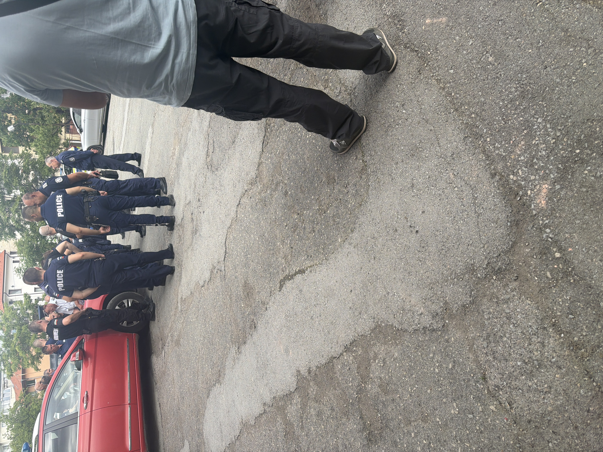 The image captures an outdoor scene in what appears to be a street or parking area in Shumen, Bulgaria, during the daytime. The foreground is dominated by the lower body of a person, seen from behind, wearing a light blue t-shirt, black cargo pants, and dark sneakers, seemingly observing the activity ahead.
In the mid-ground, a group of at least six to seven men in dark blue police uniforms are gathered. Their uniforms clearly display the word "POLICE" in white capital letters on their backs. They are standing in a loose formation or rows, facing generally towards the left side of the frame. Some individuals among the group appear to be civilians, distinguishable by their lighter colored collared shirts. One police officer is seen leaning against a red sedan, which is partially visible on the left side of the frame and appears to be a police vehicle due to a blue light visible on its roof.
Further in the background, a silver-colored sedan is parked, and residential buildings with green trees are visible, suggesting an urban or suburban setting. The ground is paved with asphalt, showing some cracks and repairs. The lighting indicates a bright day, possibly sunny or overcast, with no harsh shadows, suggesting it could be mid-morning or late afternoon. The overall atmosphere appears to be a professional gathering or briefing of law enforcement personnel.