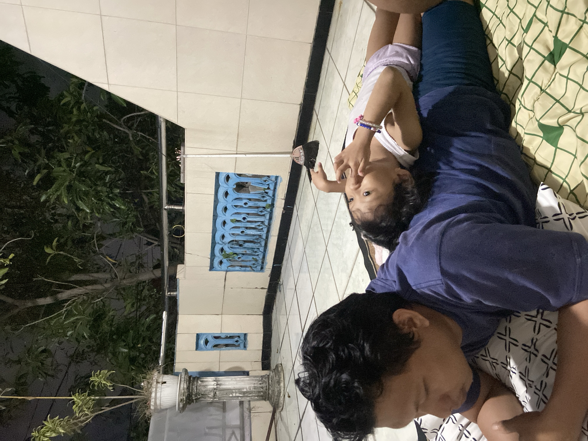 A man in a navy shirt is lying on his back on a bed with a green and white patterned blanket. A young child with curly hair, wearing a white top and lavender shorts, is lying on top of him. The child is looking upwards. They appear to be indoors, with a tiled floor visible. Through a window, there is a view of trees against a night sky. A decorative planter is visible outside the window. This is happening in Gelam, Indonesia.