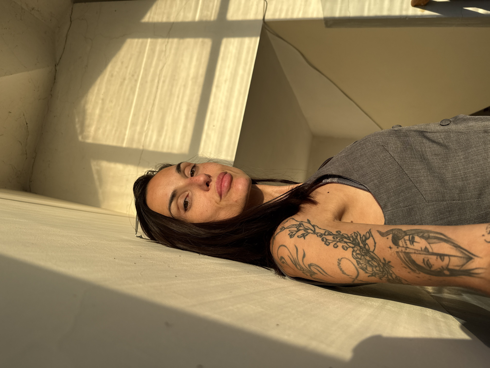 A woman is lying down indoors, in a room with sunlight streaming through a window creating a stark contrast of light and shadow on the walls. She has dark hair and a tattoo on her left arm. The woman is wearing a gray top. The location is indoors and the geographic location is unidentifiable within the image.