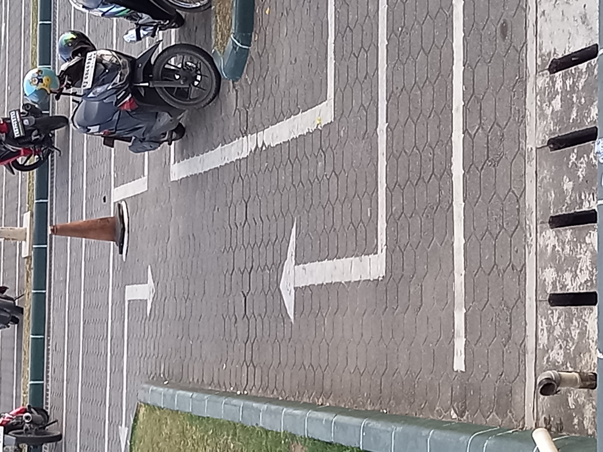 The image shows a paved area with hexagonal bricks, likely a parking lot or walkway. White arrows painted on the ground indicate direction. In the foreground, a drainage grate is visible. In the background, several motorcycles are parked. One motorcycle in the center has two helmets on it, one light blue with cartoon characters and another dark green. The scene appears to be in a public outdoor space.