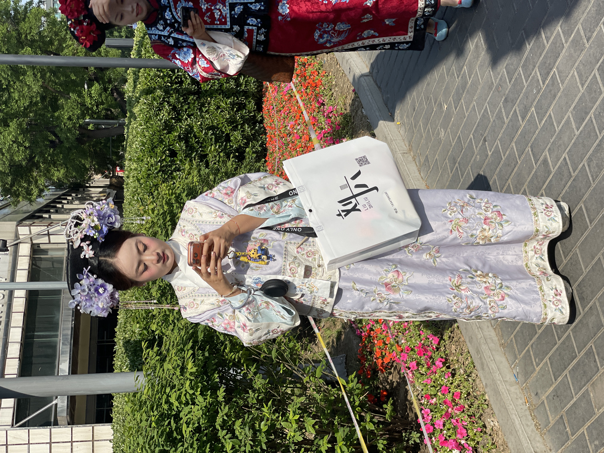 The image shows two people in traditional Chinese clothing in an outdoor setting. The primary subject is a person adorned with a purple floral headpiece, wearing a lavender floral-patterned robe, and holding a mobile phone. The individual is positioned near a flower bed with red and pink flowers and stands on a brick-paved surface. A shopping bag with Chinese characters is visible, slung over their shoulder, with "ONLY ONE" printed on the strap. The second person, only partially visible in the background, is wearing a red and blue traditional outfit. The environment includes greenery, trees, and part of a building with windows. The scene appears to be set in an urban area with manicured gardens. The location is Dongcheng District, China.