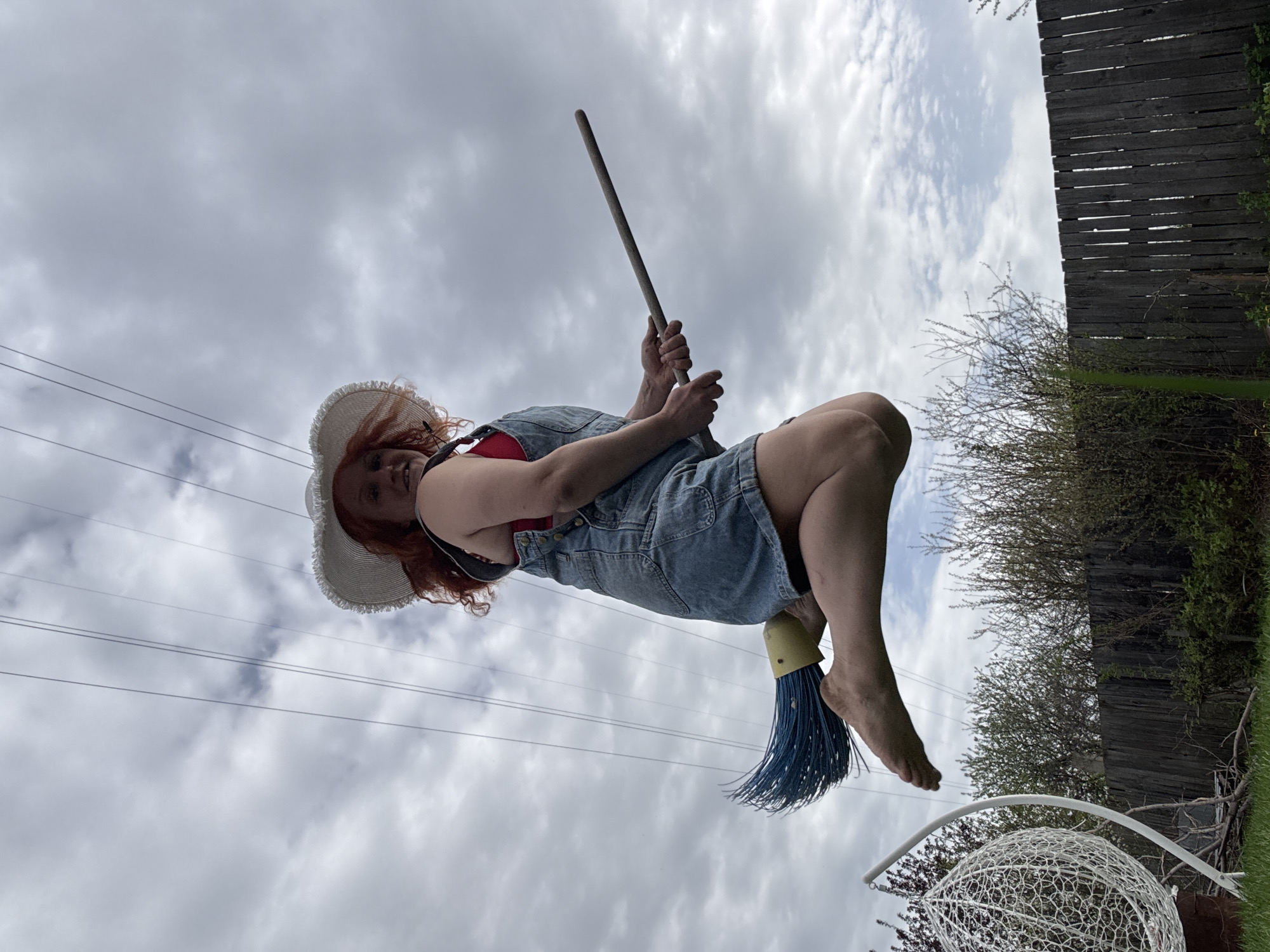 A woman is airborne, appearing to ride a blue push broom. She's wearing a denim overall dress, a red top, and a white straw hat. She's holding the broom handle with both hands. The backdrop consists of a cloudy sky, a wooden fence, trees, and power lines. There's also a white outdoor chair or structure visible at the bottom of the frame. The location is speculated to be Chaikovsky, Russia.