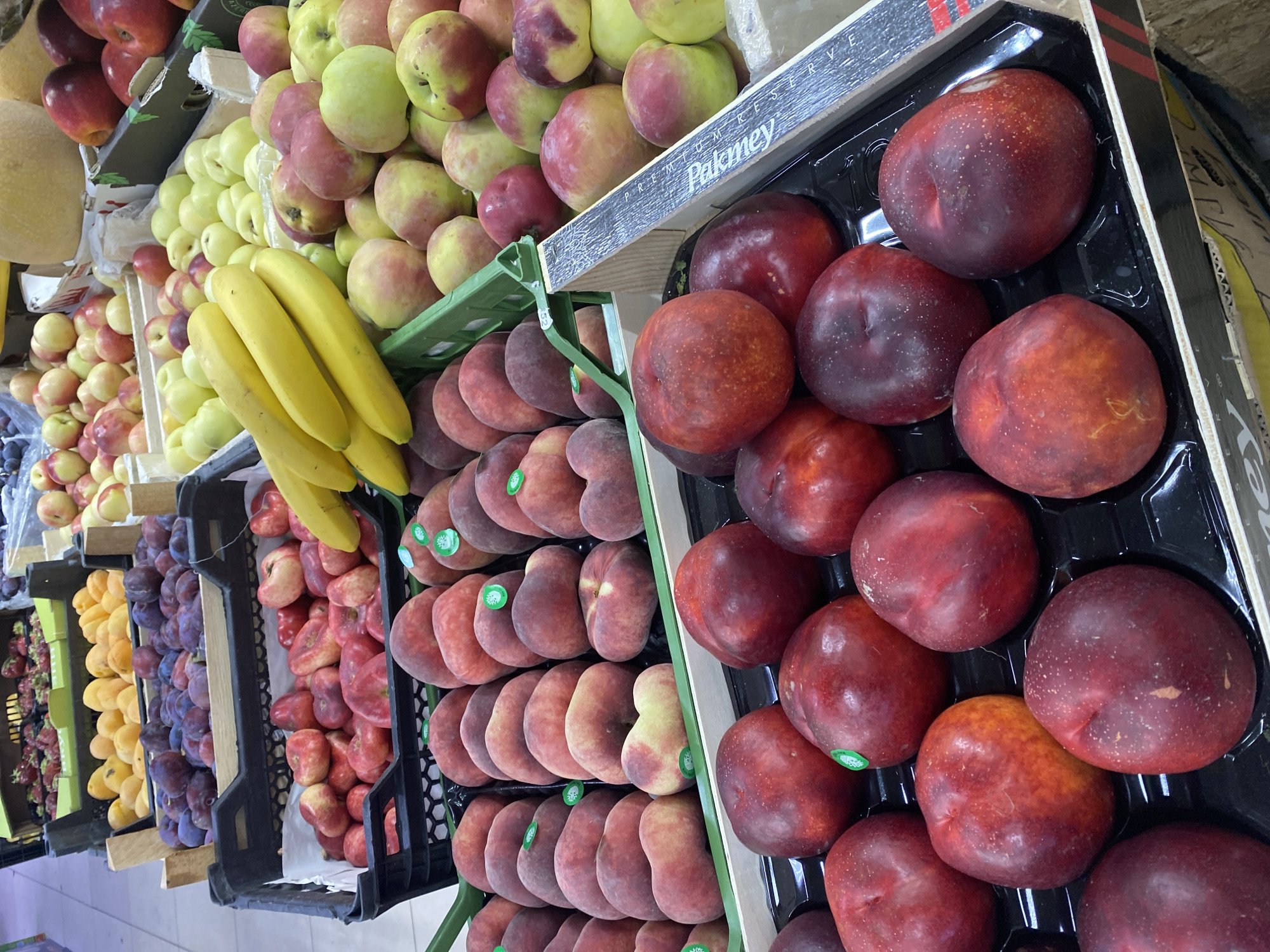 The image depicts a vibrant display of fresh fruits in what appears to be a market or grocery store. In the foreground, several crates are filled with various types of produce. Prominently featured are ripe peaches and nectarines, with their characteristic red and yellow hues. Some of these fruits have small green stickers on them. Behind these, there are bunches of yellow bananas. Further back, crates overflow with plums, apricots, and apples in shades of green and red. In the background, a large, pale yellow melon is visible. The scene suggests a bustling retail environment, likely a section of a supermarket or a dedicated fruit stall. The lighting indicates an indoor setting, possibly with artificial lights illuminating the produce. There are no people visible in the frame, and the focus is solely on the arrangement and variety of the fruits. The presence of price tags or labels is not clearly discernible in this image. Based on the assumption that this image was taken in Moscow, Russia, it is likely a scene from a large grocery store or a farmers' market within the city, offering a wide selection of seasonal fruits to customers.