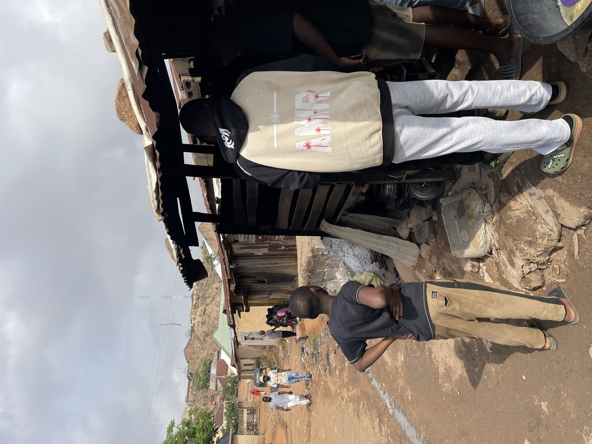 The image depicts a scene in a densely populated area in Jos, Nigeria. The foreground shows two young men: one wearing a black t-shirt and light brown trousers with dark side panels, standing with arms crossed; the other wearing a beige and black jacket, gray sweatpants, and green Crocs. They are positioned near a dilapidated structure with a corrugated metal roof and rough wooden walls. Other people are visible in the distance, walking along a dirt road lined with buildings of varying condition. A rocky hill dominates the background, and a communication tower is visible against a cloudy sky. The environment appears impoverished, with evidence of disrepair and limited infrastructure.