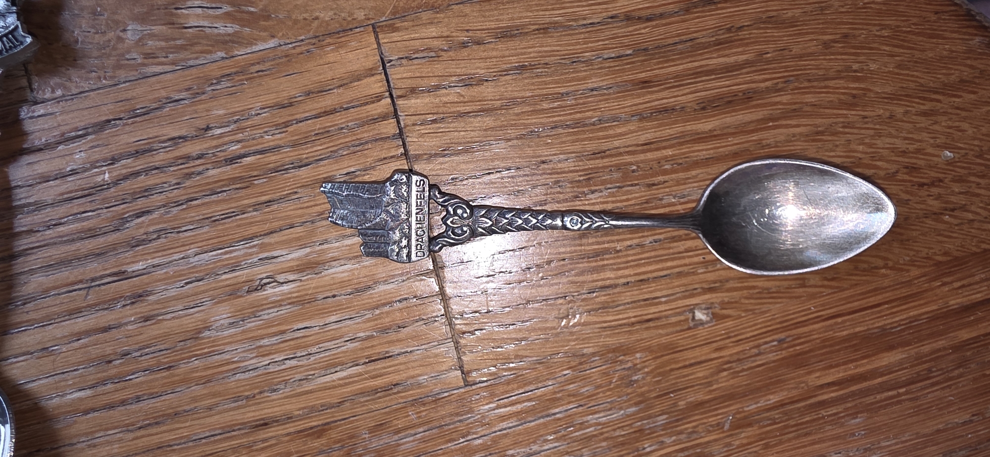 An ornate silver spoon with a miniature castle carving on its handle, labeled "DRACHENFELS", rests on a wooden table. The spoon is positioned vertically with the bowl facing downwards and slightly to the left. The table surface displays a prominent wood grain pattern with visible lines and texture.