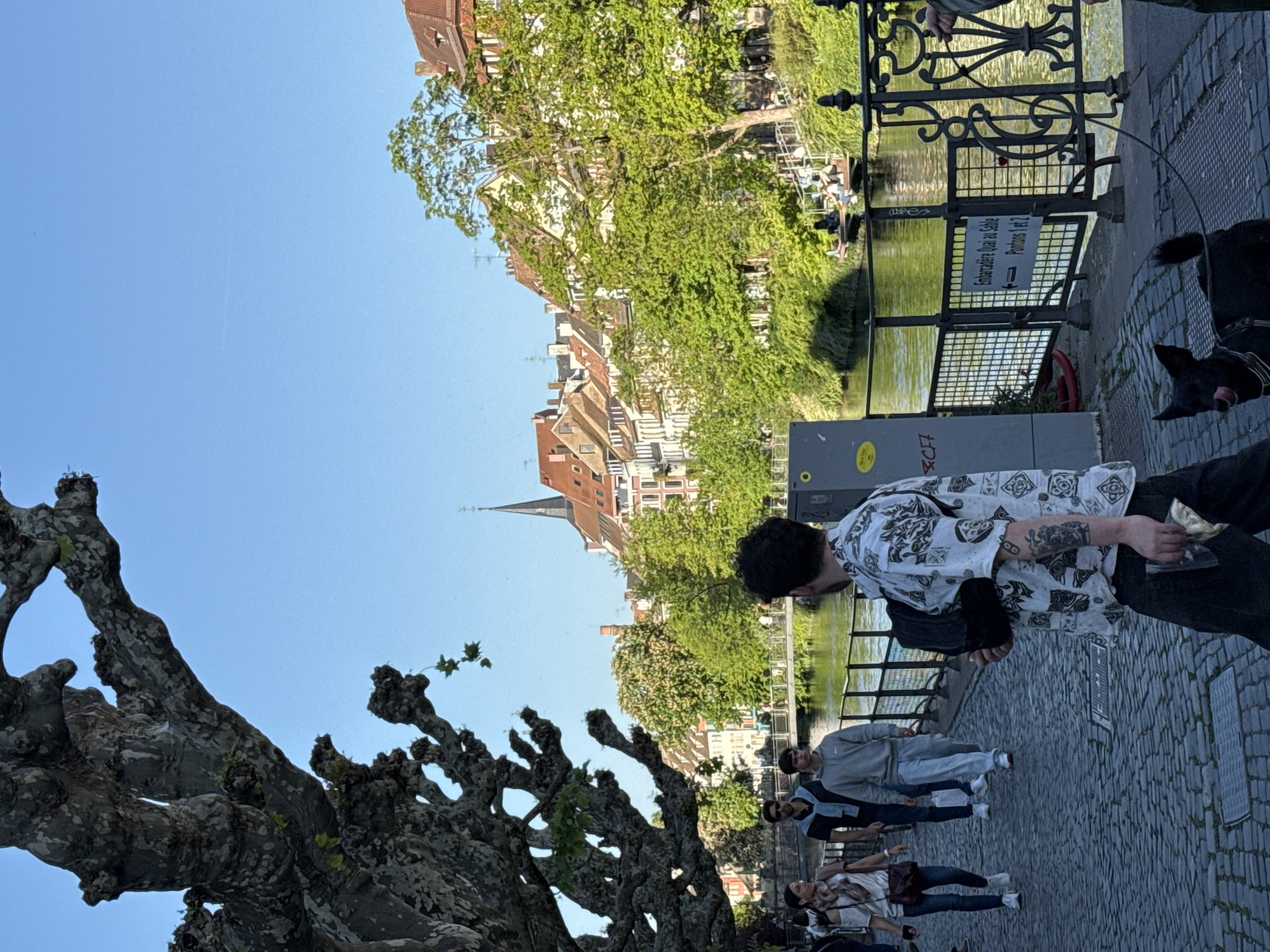 Here is a description of the image:
The photo depicts a street scene in Strasbourg, France, specifically along the Quai au Sable, next to the river. The photo is taken from a slightly elevated angle. A large, bare tree with thick branches dominates the left foreground. Along the river, there are pedestrians walking, including a man with tattoos wearing a patterned shirt standing next to the railing of the quay, and a black dog at his side. Other pedestrians, including a group of people walking together, can be seen in the middle ground. In the background are buildings with orange roofs and the spire of a church. The waterway is lined with greenery and there is some seating visible on the opposite bank. A sign with text in French referencing the "Embarcadere Quai au Sable" is attached to the railing. The sky is blue and clear.