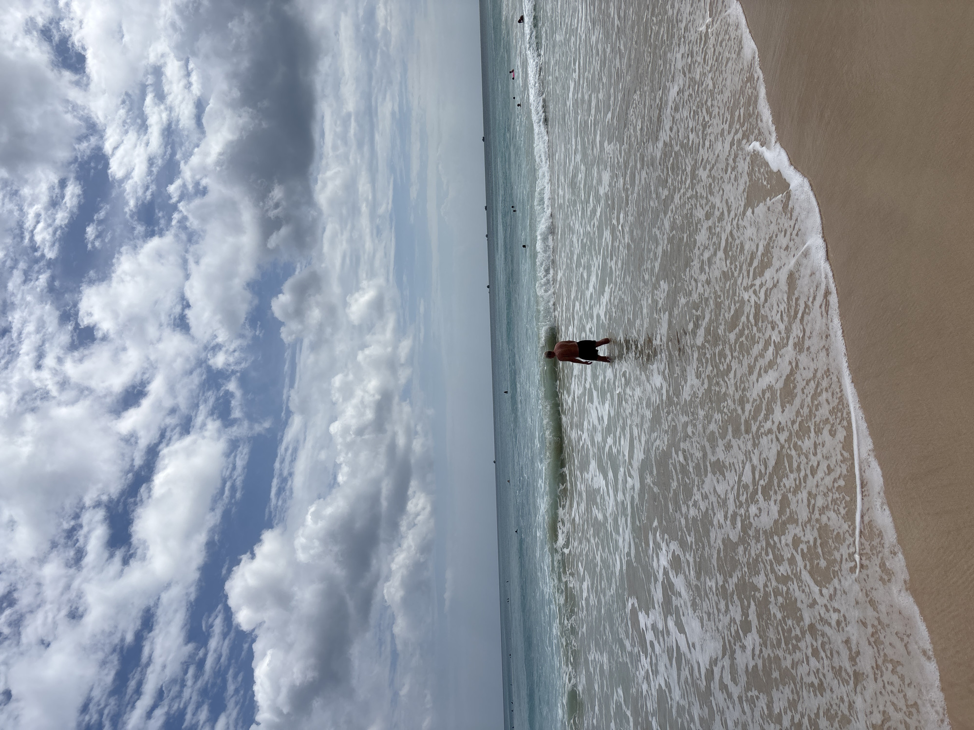 A man stands waist-deep in the turquoise ocean on a sandy beach, facing away from the camera. He is wearing dark shorts. Waves with white foam are breaking around him and receding towards the shore. In the distance, several boats are visible on the horizon, and other people are swimming and playing in the water. The sky above is a vibrant blue, filled with scattered white and gray clouds, suggesting a partly cloudy day. The image appears to have been taken in Kuta Selatan, Indonesia, a popular tourist destination known for its beaches and surf.