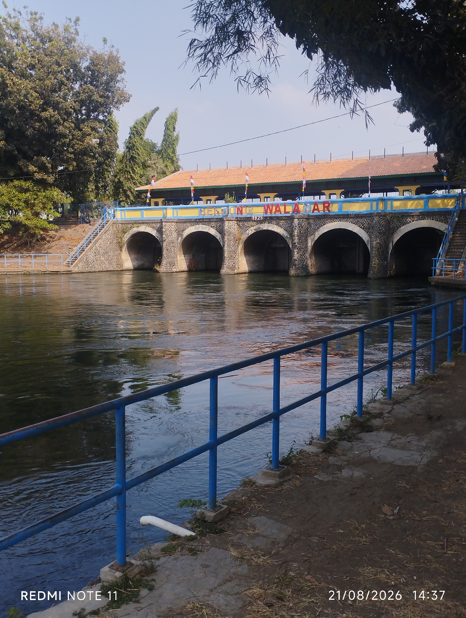This is an outdoor, ground-level, medium shot of a water dam with arches and a building on top, situated in the city of Karawang, Indonesia. The dam has a stone facade and is spanned by a bridge painted in yellow and blue, with the words "BENDUNG WALAHAR" visible on it, along with the year "1925". Small flags in red, white, yellow, and blue are attached to the bridge railing. To the left of the dam, there are stairs leading up to the bridge, and on the far left, a large tree with dense foliage stands. The water flowing through the dam is dark and reflects the sky and the structure. A blue metal railing lines the edge of the pathway on the right side of the image, where the ground is covered in dirt and some dried leaves. A white PVC pipe is visible near the water's edge in the lower left quadrant. The sky is clear and blue, with some tree branches visible in the upper right corner. A timestamp in the bottom right corner reads "21/08/2026 14:37", and "REDMI NOTE 11" is in the bottom left corner. There are no people or animals visible in the scene. The scene appears to be a public area, possibly a tourist attraction or a functional water management structure.