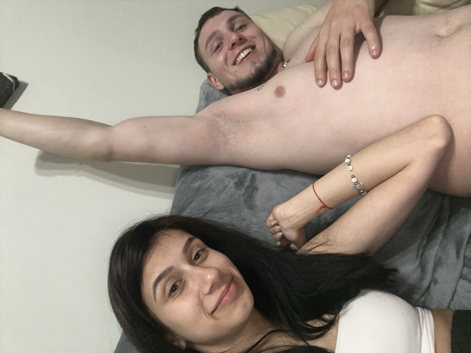 A shirtless man with a tattoo on his chest and a short beard smiles and lies on a gray couch. He is next to a woman with long dark hair and a white top. The woman is smiling slightly. There are no specific details to indicate the location as Plovdiv, Bulgaria.