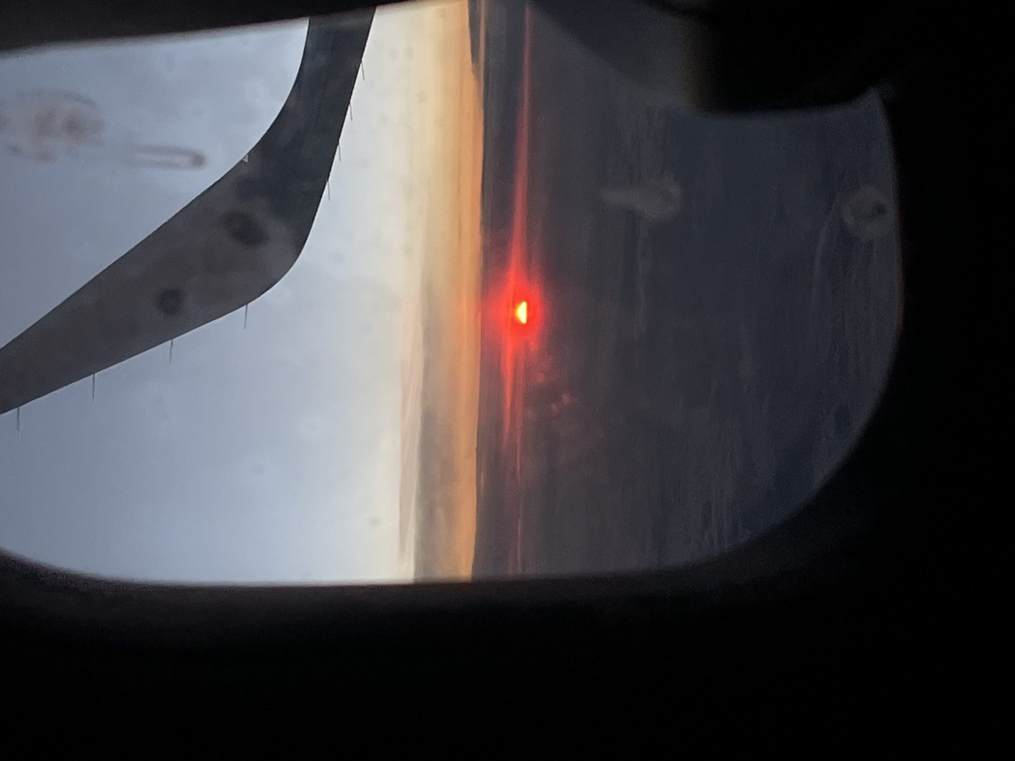 The image is taken from an airplane window, looking out at a sunset. The sun appears as a bright, small, red, semi-circular shape near the horizon, casting a warm, orange glow on the clouds below. Part of an airplane wing is visible in the foreground, obscuring the left side of the view. The scene suggests an airborne journey during twilight. The overall mood is serene, capturing the beauty of a sunset from a unique vantage point. The exact location is Novokuznetsk, Russia. There is no visible text in the image.