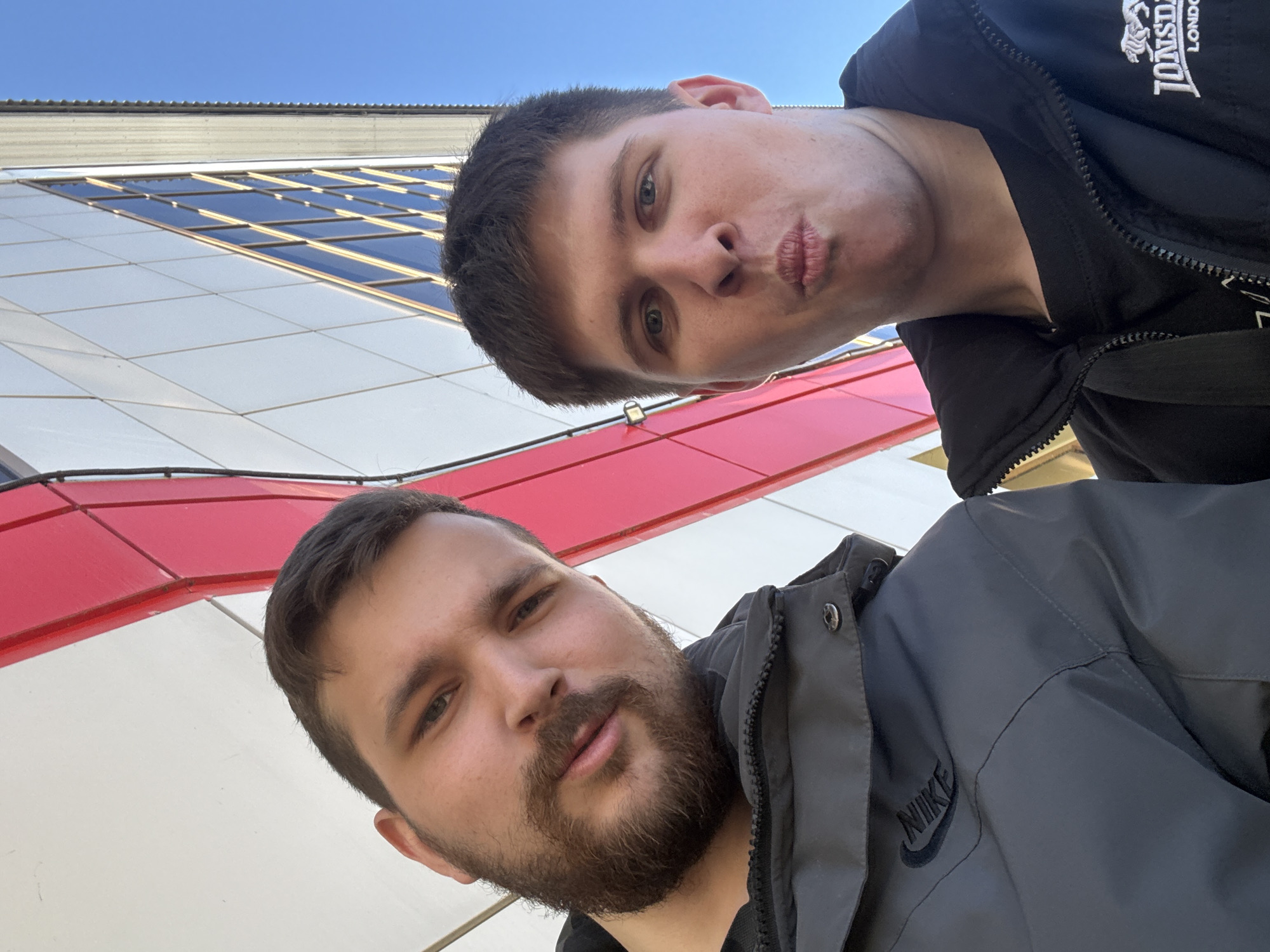 Two men take a selfie in front of a building. The building has white, red, and glass paneling. The man at the top is wearing a black jacket with a "LONSDALE LONDON" logo. The man at the bottom is wearing a gray jacket with a "NIKE" logo. The photo was taken in Chaikovsky, Russia.