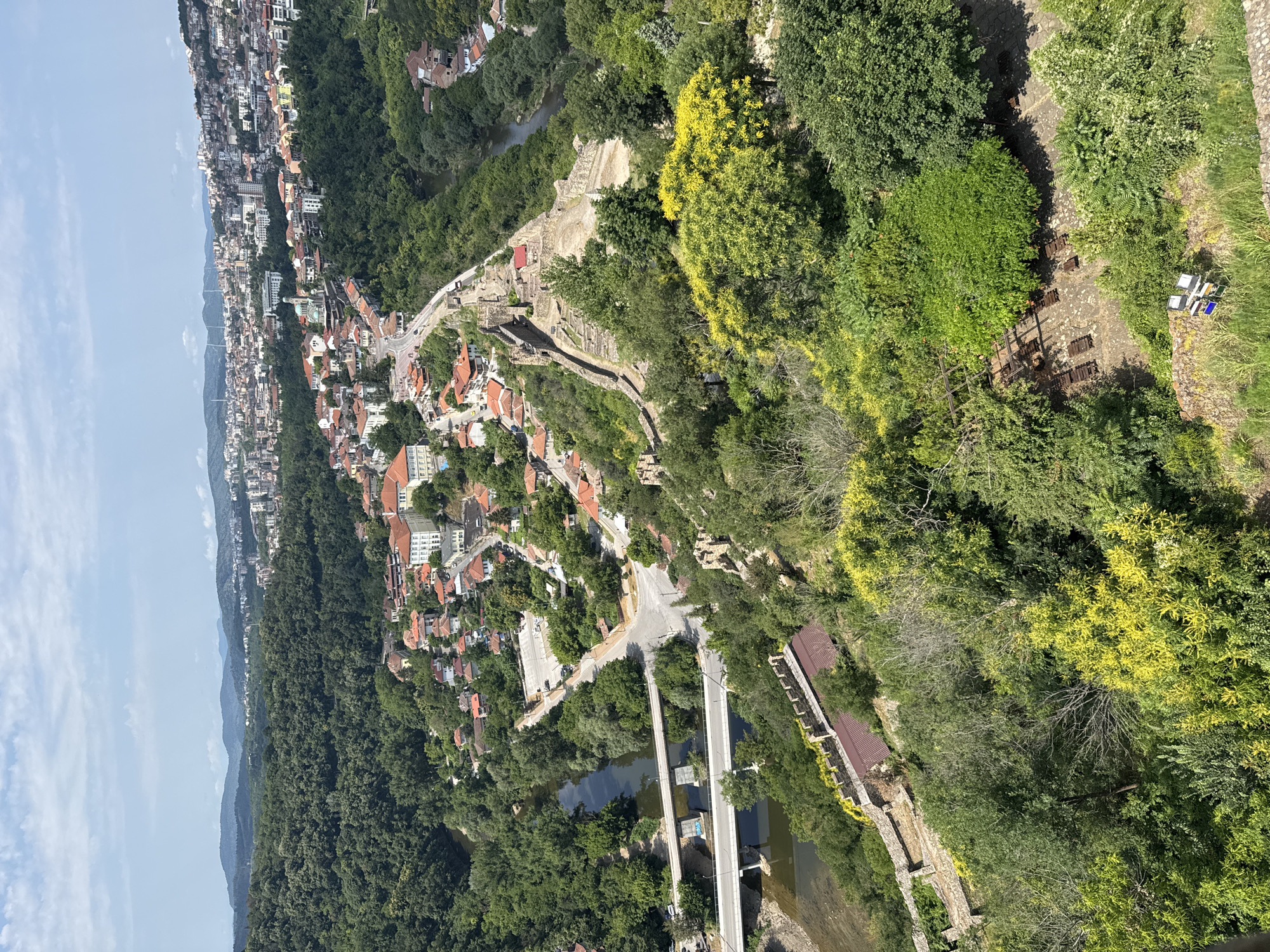 An aerial, high-angle perspective captures a panoramic view of the historic city of Veliko Tarnovo, Bulgaria, nestled within a lush, verdant landscape. The Yantra River curves through the scene, spanned by modern concrete bridges. On the left, a riverbank lined with trees leads to a road. The right side of the image is dominated by the Tsarevets fortress, its ancient stone walls and structures tracing the contours of the hill. Buildings with red-tiled roofs are clustered on the hillsides, interspersed with dense green foliage. In the distance, a sprawling urban area extends across the horizon, with more distant hills fading into the hazy blue sky. The foreground is filled with vibrant green trees and shrubs, with a prominent yellow-flowering bush adding a splash of color. A few modern streetlights are visible on a stone pathway in the lower right corner.