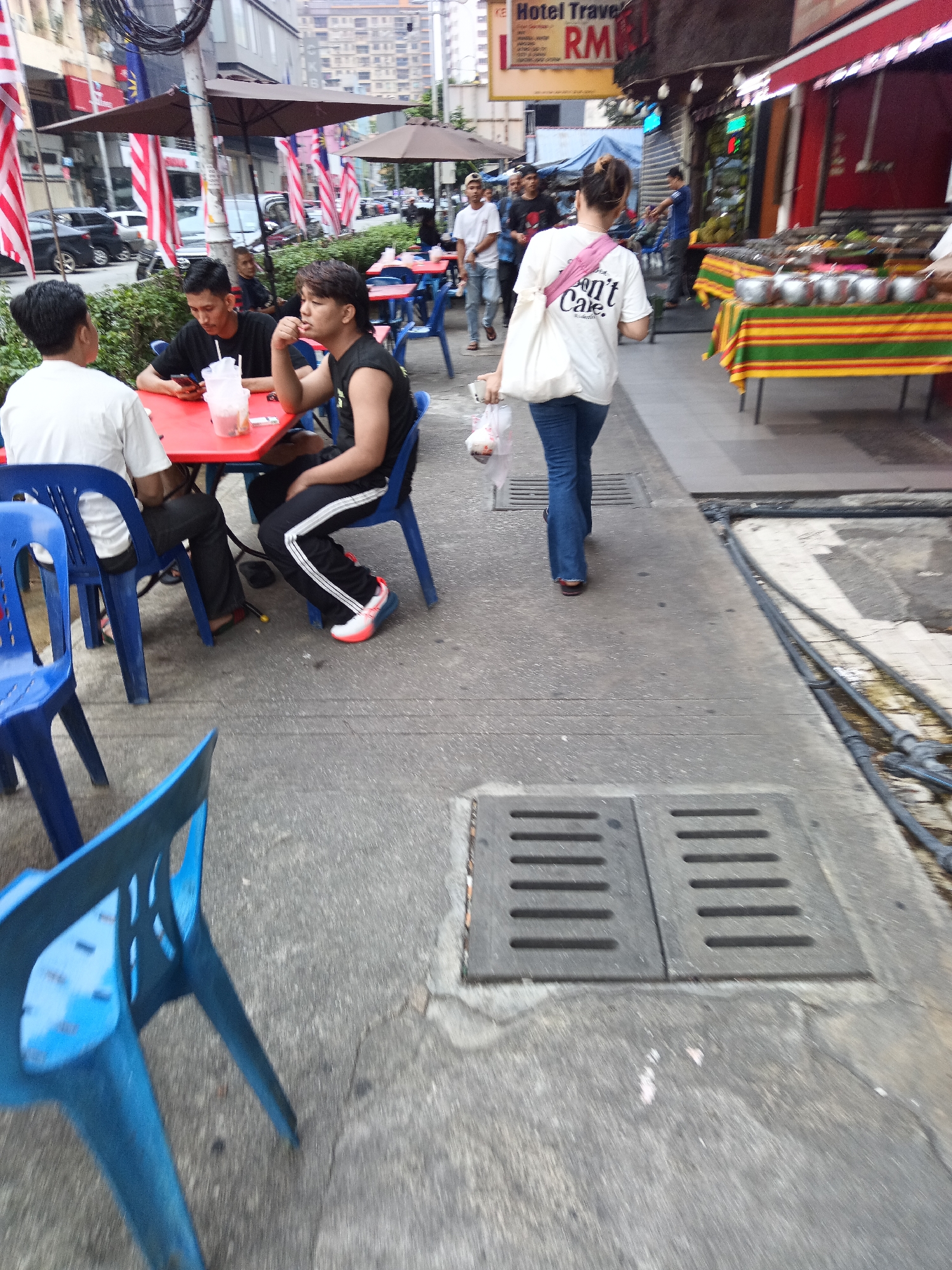A group of people are dining at outdoor tables on a street in Kuala Lumpur, Malaysia. The tables are red and set with blue plastic chairs. Two men are seated at one table, and another man is seated at a table behind them. A woman in a white t-shirt and blue jeans walks away from the camera, carrying a plastic bag. In the background, there are buildings, flags, and umbrellas. There is also a stall with food displayed on a table covered with a striped cloth. The street has manhole covers and exposed pipes.