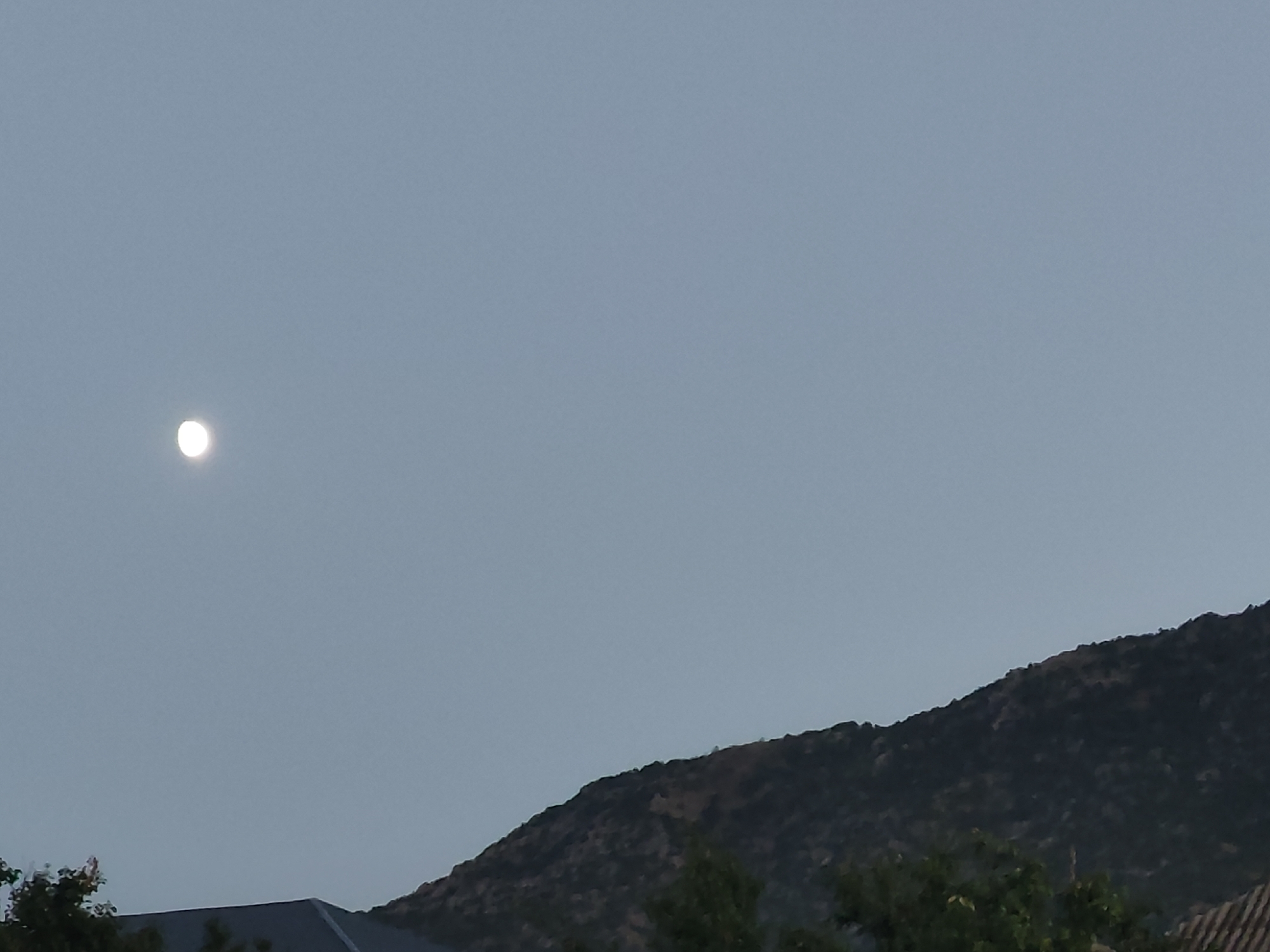 The image depicts a hazy, pale blue sky with a small, bright moon visible in the upper left quadrant. Below the sky, a dark, undulating mountain range occupies the lower right portion of the frame. The mountain's surface is textured and appears to be covered in scrub or sparse vegetation. In the lower foreground, the dark roofs of several buildings are partially visible, along with some greenery, suggesting a semi-rural or suburban setting. The lighting is dim, indicative of either twilight or an overcast day. The scene appears to be a landscape, with no people or discernible activity. The specific location in Leninkent, Russia, is not visually identifiable from the image itself.