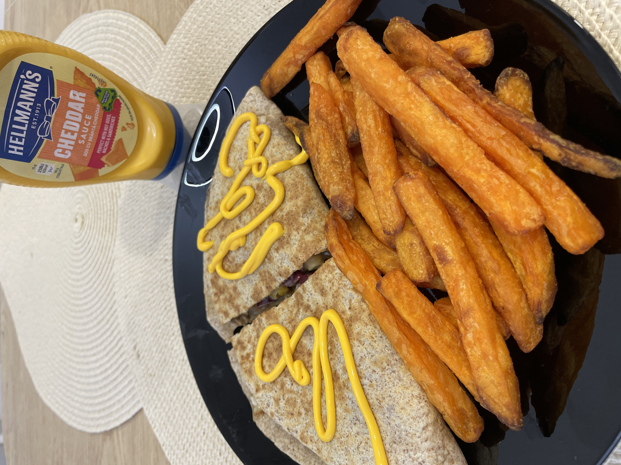 The image shows a black plate containing two cut quesadillas drizzled with yellow cheddar sauce and a pile of orange fries. A bottle of Hellmann's Cheddar Sauce is placed near the plate. The plate is resting on a round, woven placemat that is on a light-colored wooden surface.