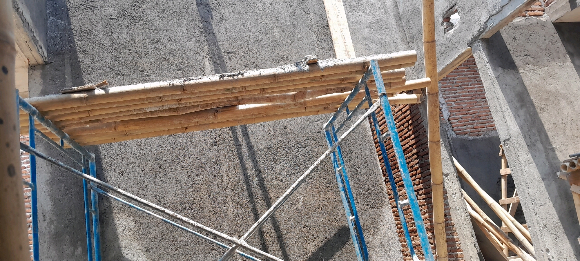 The image depicts a construction site in Karangduren, Indonesia, with a focus on bamboo scaffolding supporting a platform. The scaffolding, primarily made of bamboo poles bound together, is positioned against a rough concrete wall. A section of the wall shows exposed brickwork. The platform itself is constructed from parallel bamboo poles, some of which appear to be reinforced with thin pieces of wood. The scene is bathed in sunlight, casting shadows across the textured concrete. The overall impression is that of an unfinished building undergoing construction, with a clear emphasis on the use of traditional scaffolding materials. There are no people visible in the image.